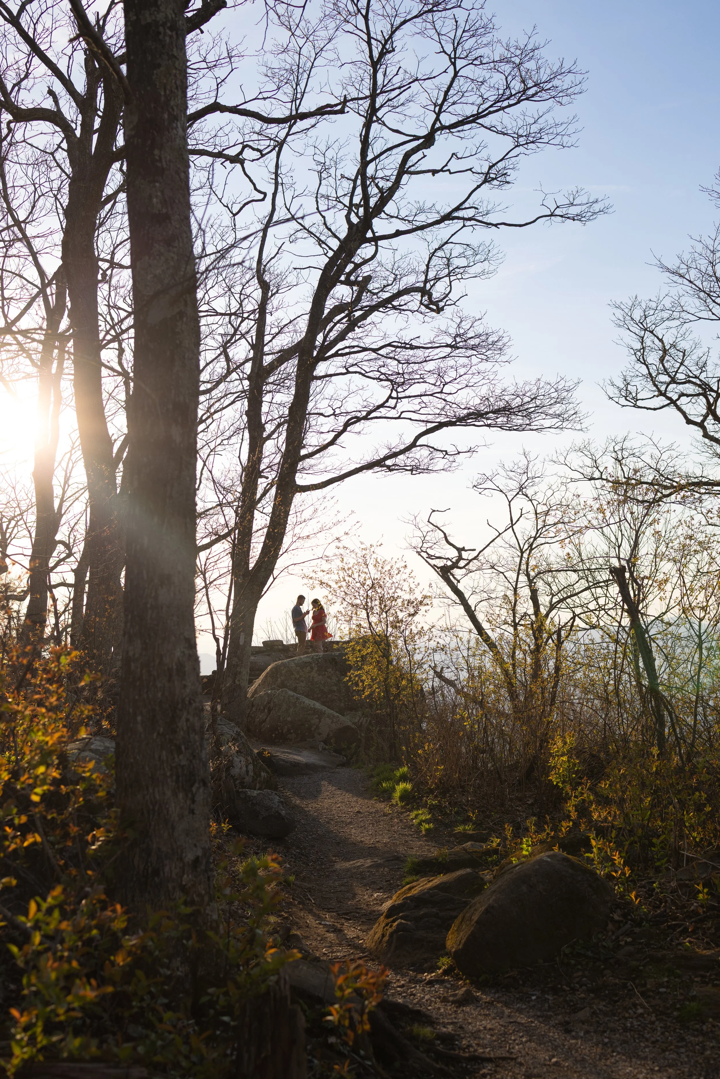 A couple standing on a rocky trail in a wooded area with leafless trees, sunlight peeking through, and a clear blue sky.