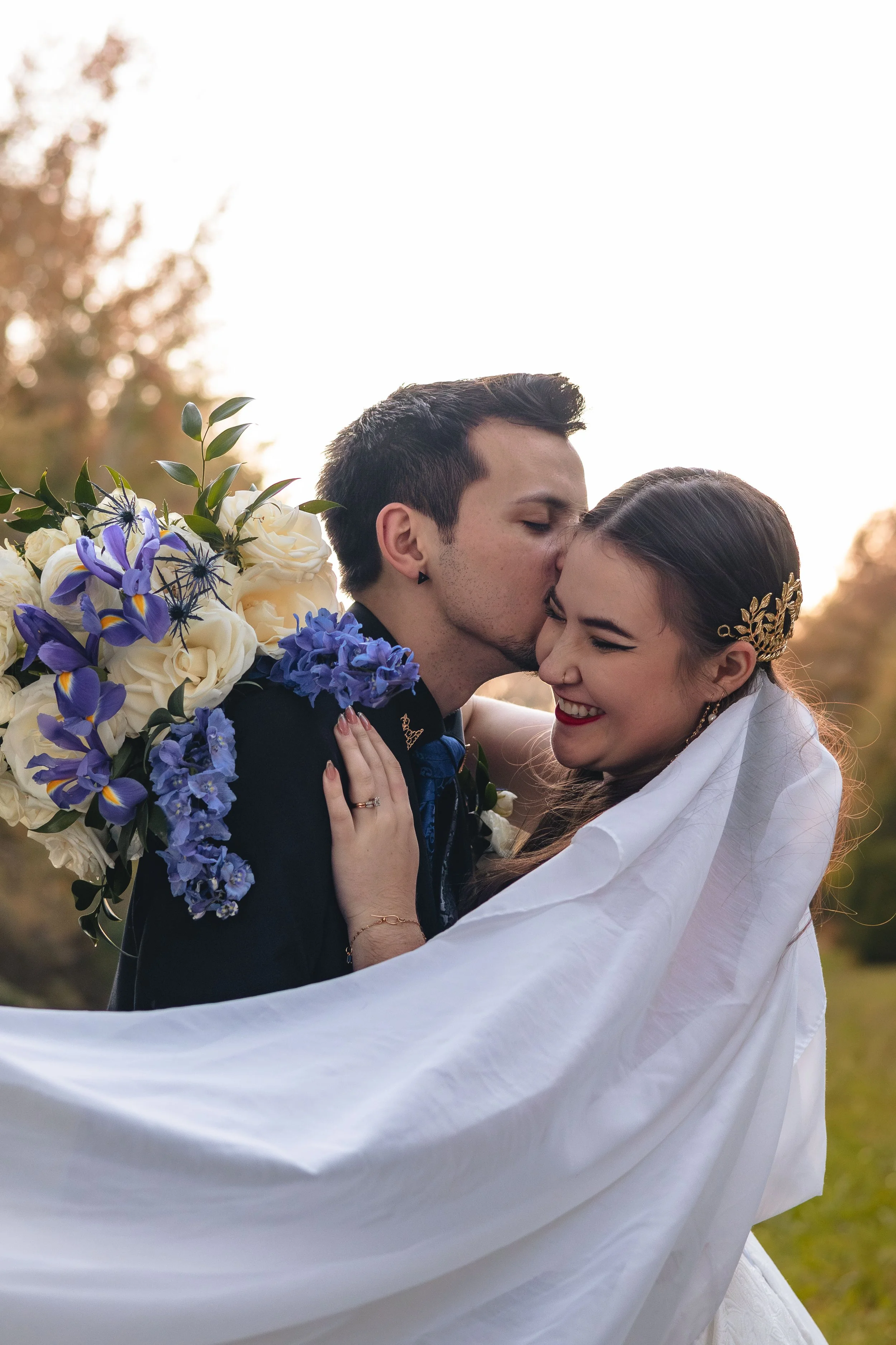 A wedding couple hugging outdoors during sunset, with a large bouquet of white and purple flowers on the bride's back.
