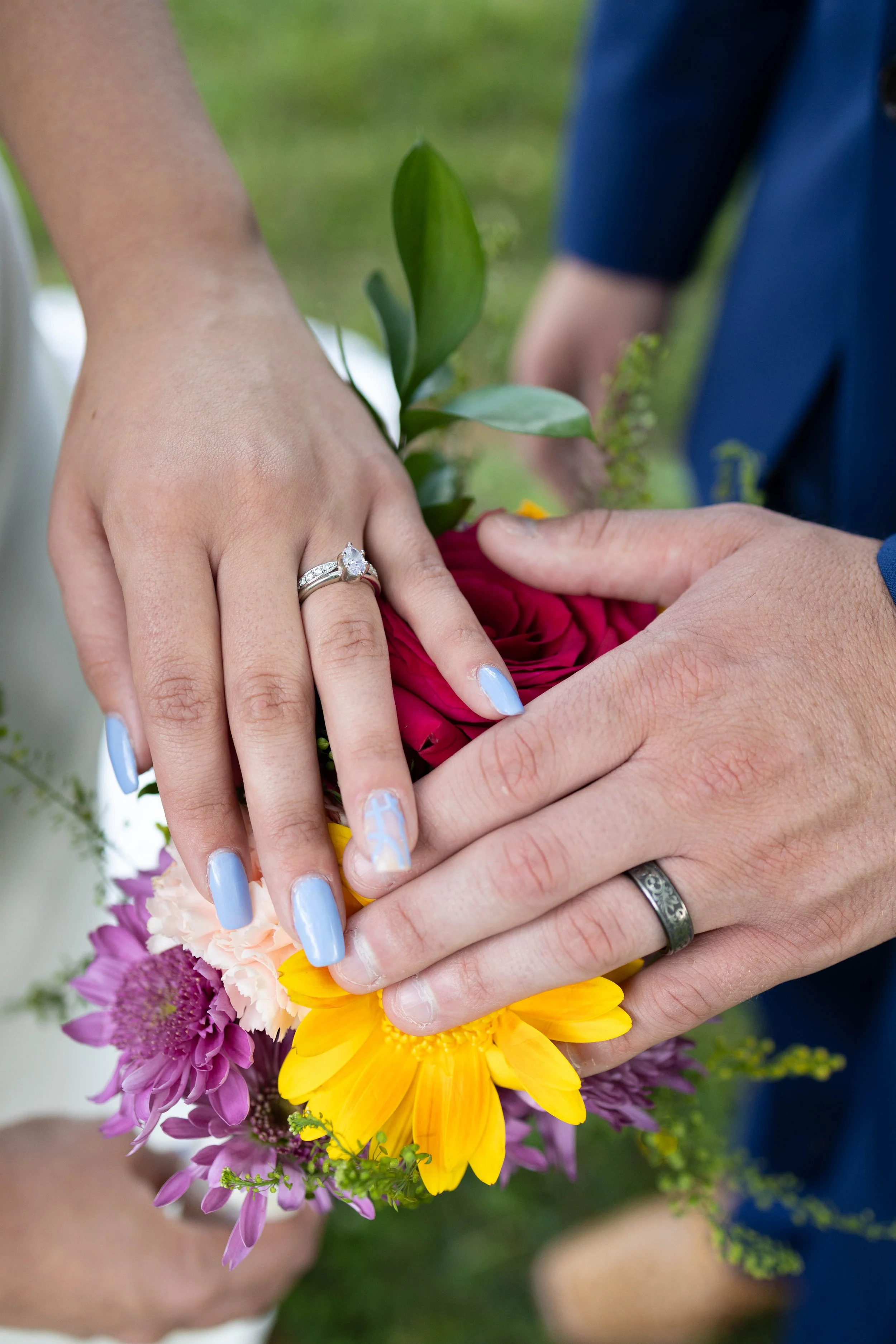 Two people are holding a bouquet of colorful flowers together. One person's hand displays an engagement ring with a large central diamond. The person's nails are painted light blue with white accents. The scene appears to be outdoors.