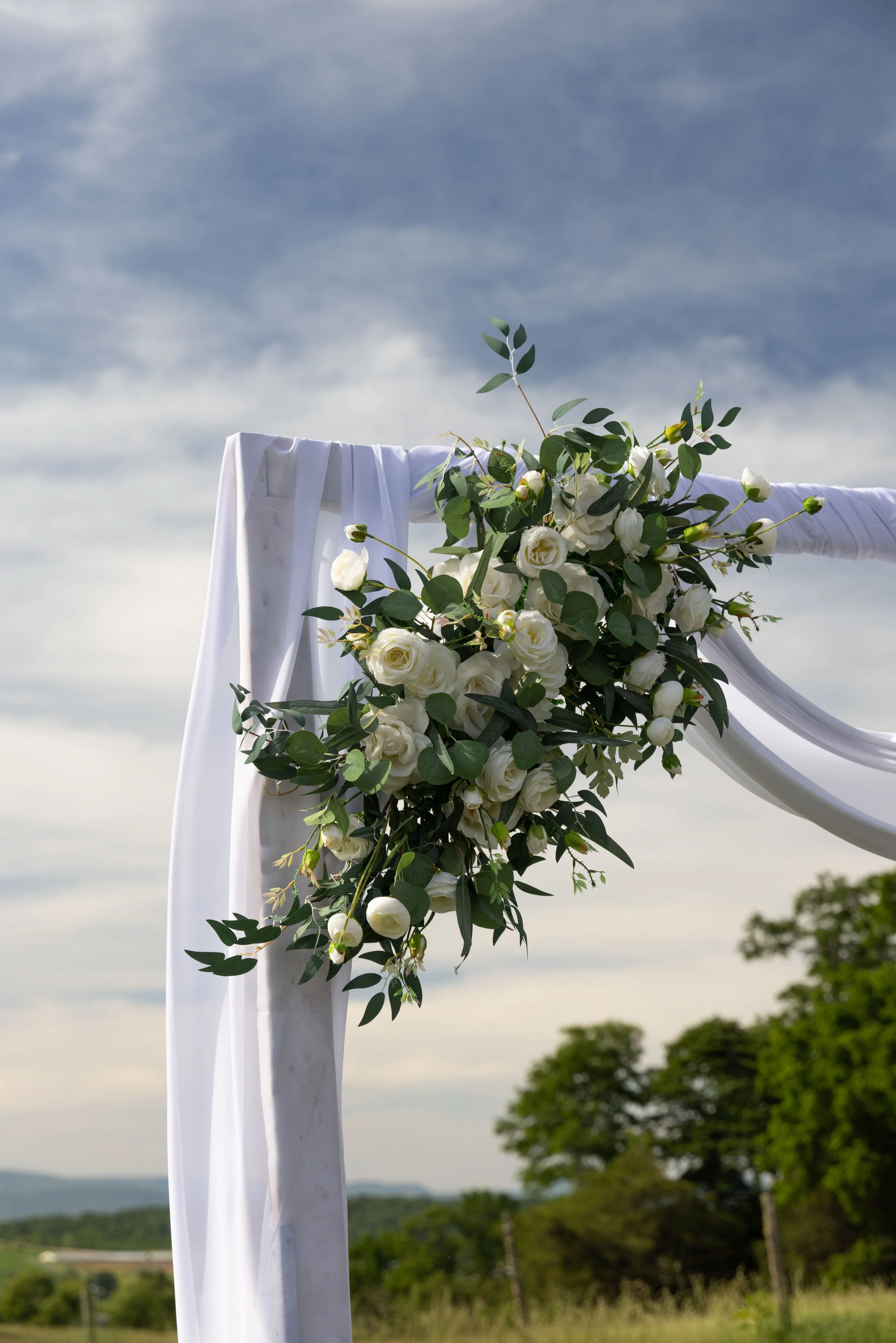 White fabric draped over a pole with a large bouquet of white roses and green leaves, set against a cloudy sky with trees in the background.
