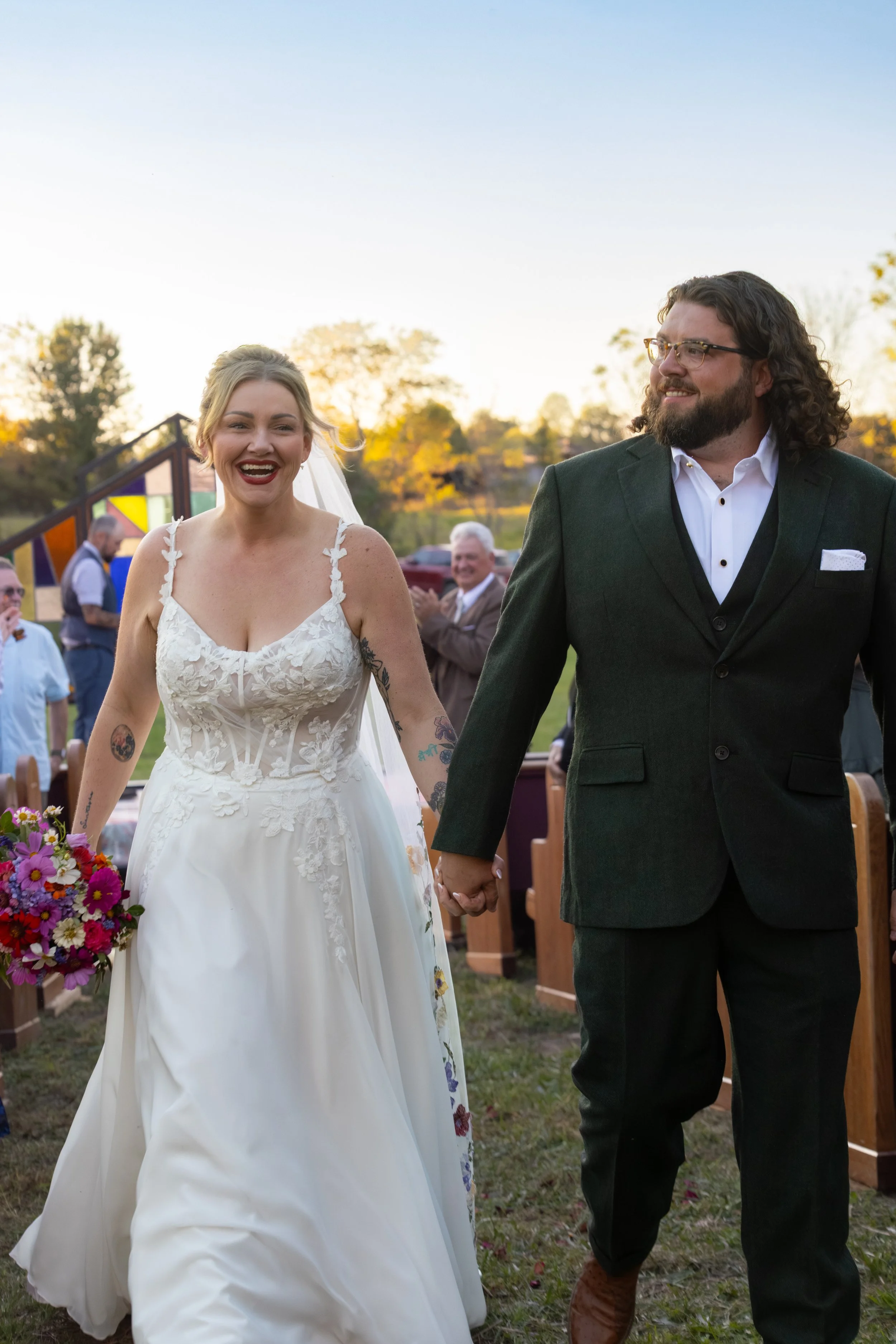 A bride and groom holding hands and walking outdoors during their wedding ceremony at sunset. The bride wears a white lace wedding dress, and the groom is dressed in a dark green suit with a white shirt. Guests are clapping in the background.