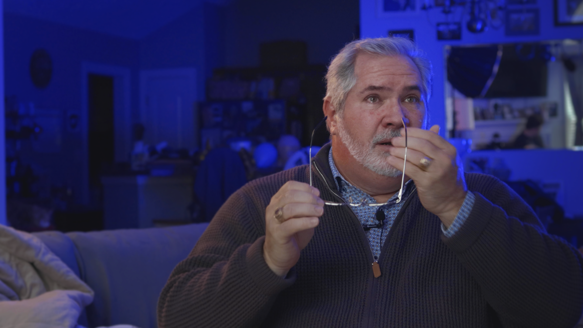 An older man with gray hair and a beard sitting on a couch, holding glasses in his hand, with a surprised or shocked expression on his face. He appears to be in a dimly lit room with blue lighting.