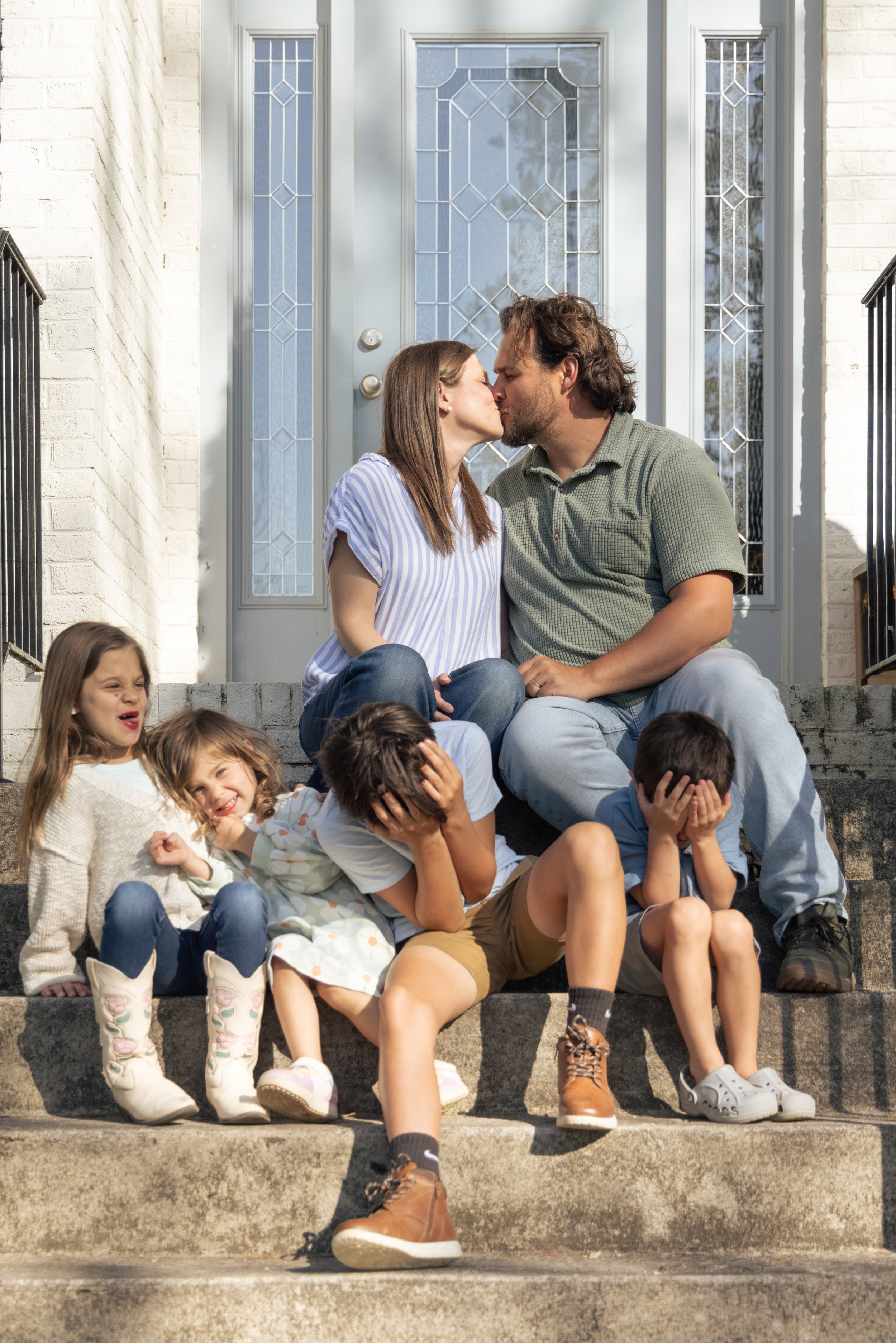 Family sitting on front steps of house, two adults kissing, children laughing, playful, casual clothing, sunny day.