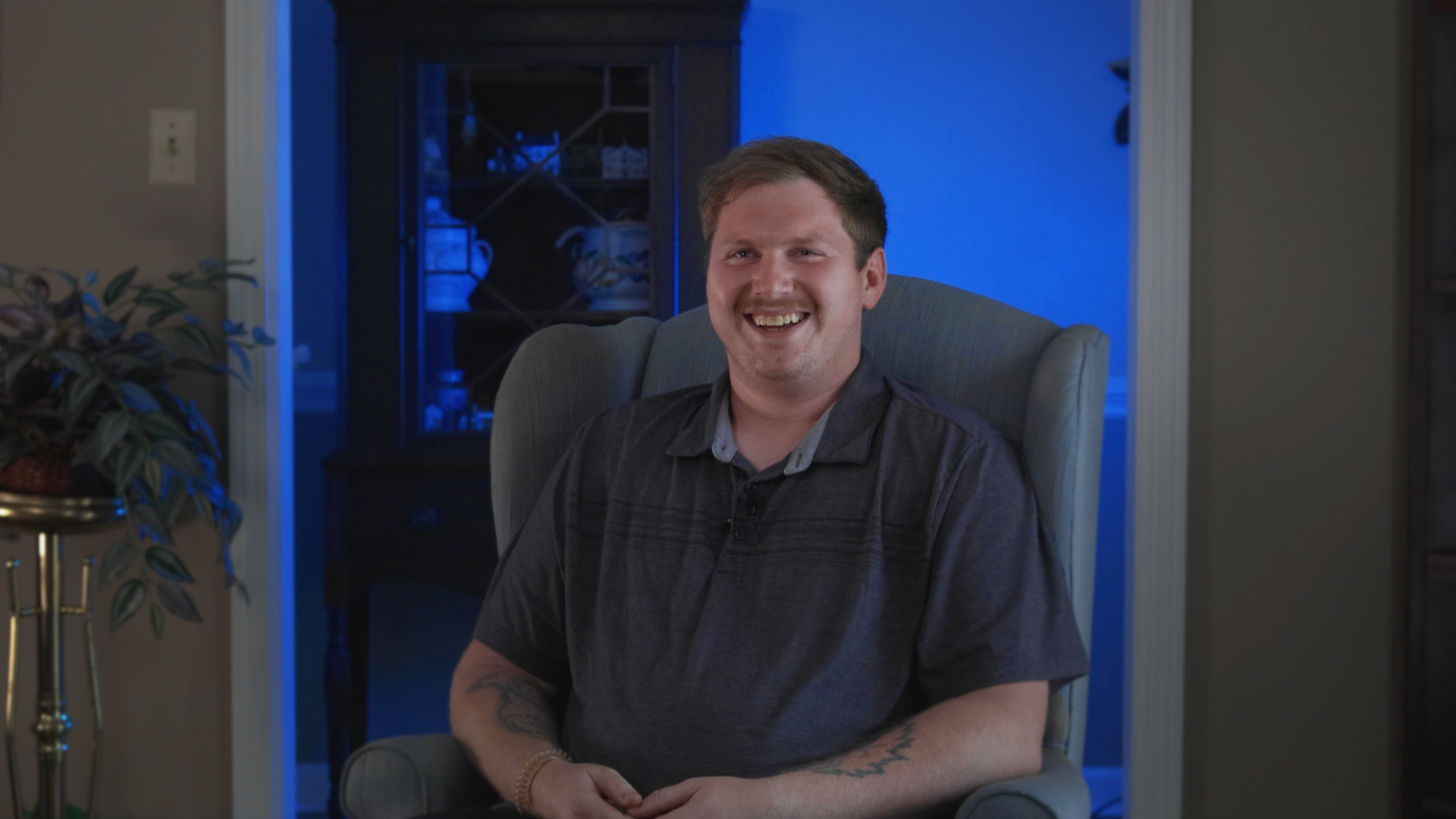 A man with short brown hair and a mustache, smiling while sitting in a beige armchair indoors. Behind him, there is a blue-lit room with a dark wooden cabinet displaying dishes.