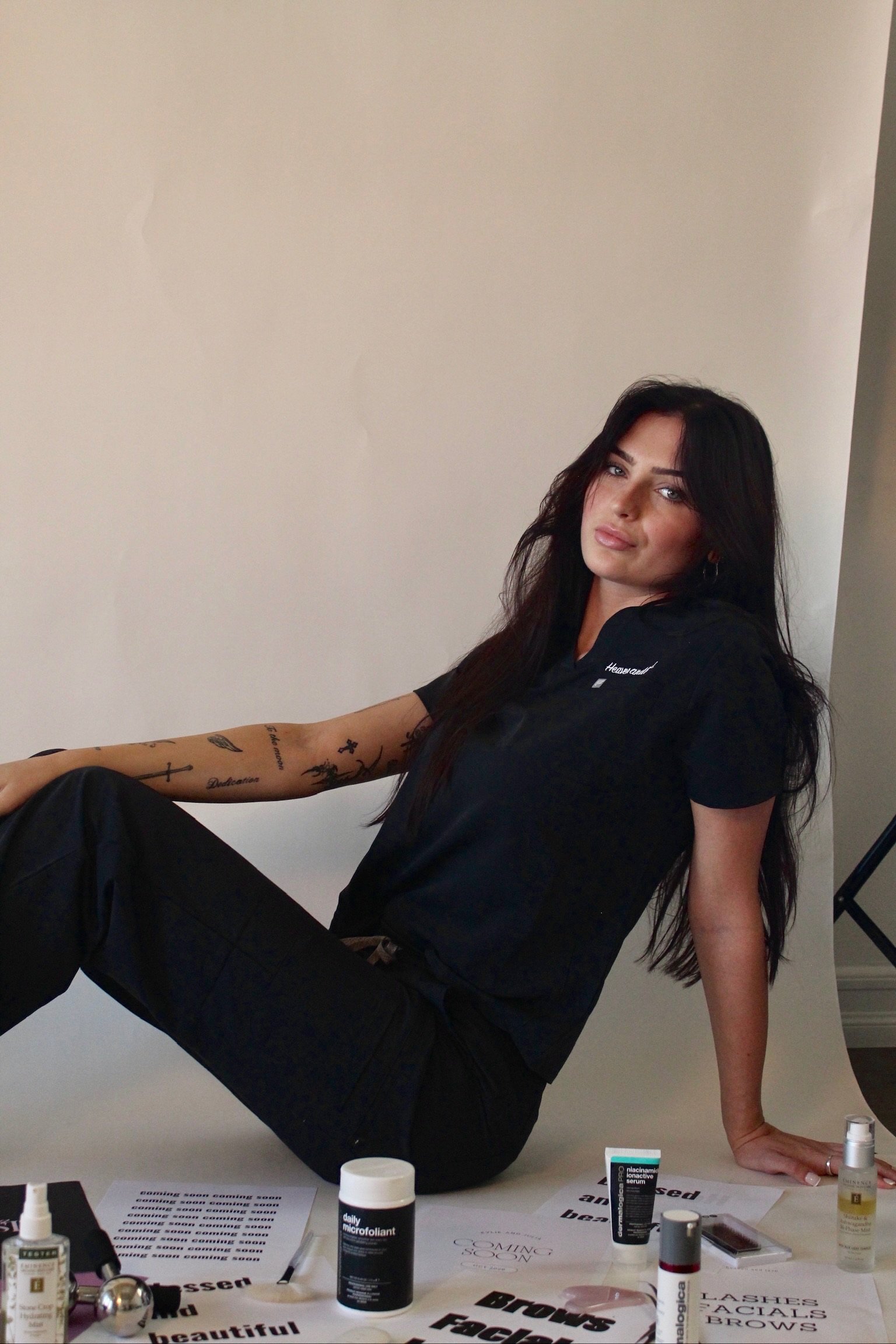 A young woman with long dark hair and tattoos on her arm, sitting on the floor in front of a white background, surrounded by skincare products and papers with black and white text.