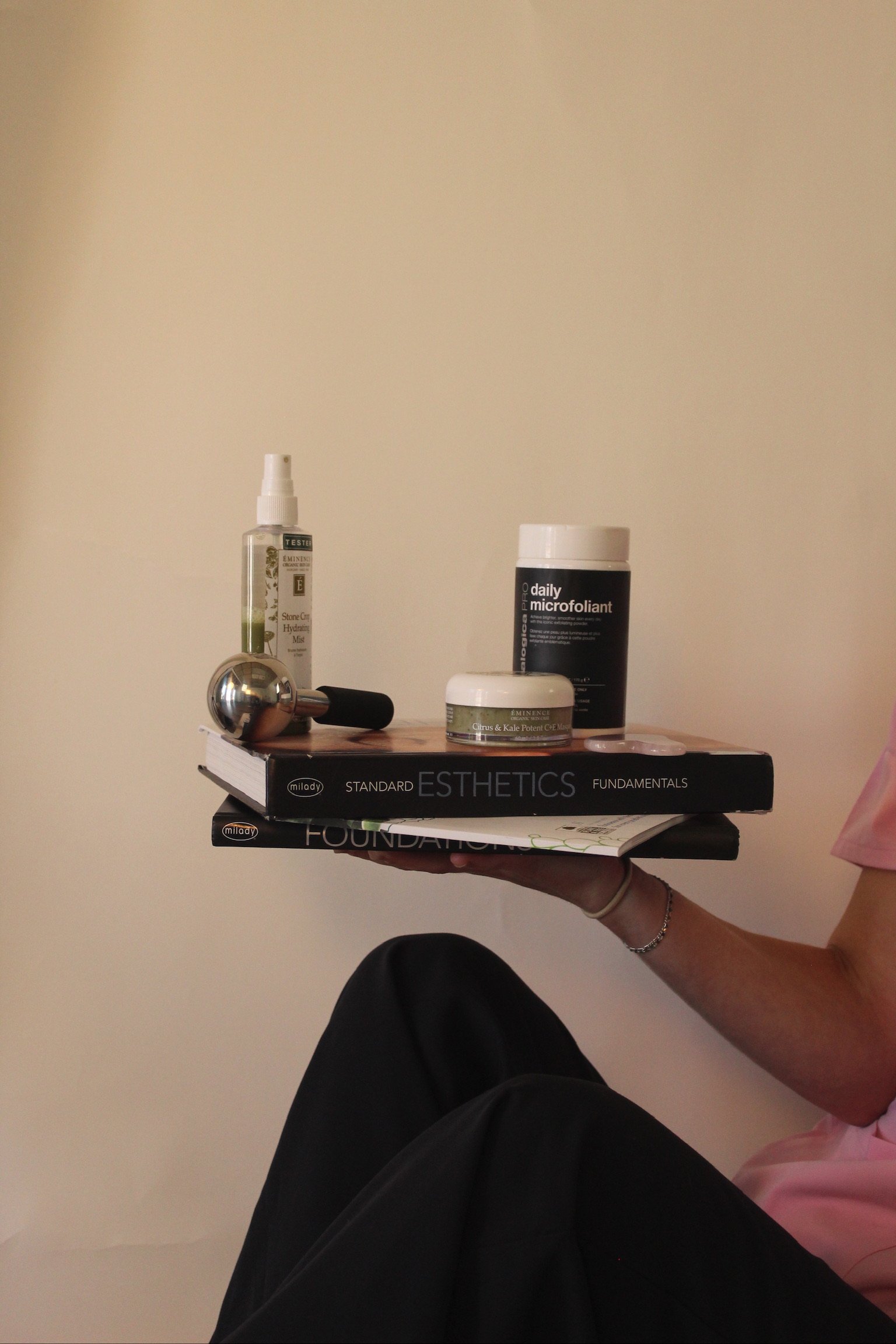 Person holding a stack of books, skincare products, and a facial roller against a plain wall.