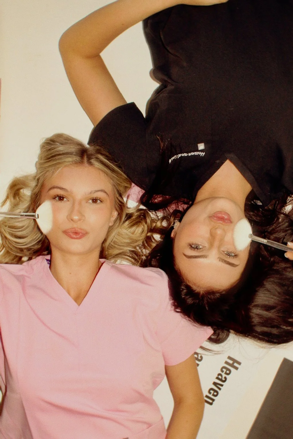 Two women lying on the floor, one wearing a pink outfit and the other in a black outfit, both holding makeup brushes to their cheeks, with hair spread around them.