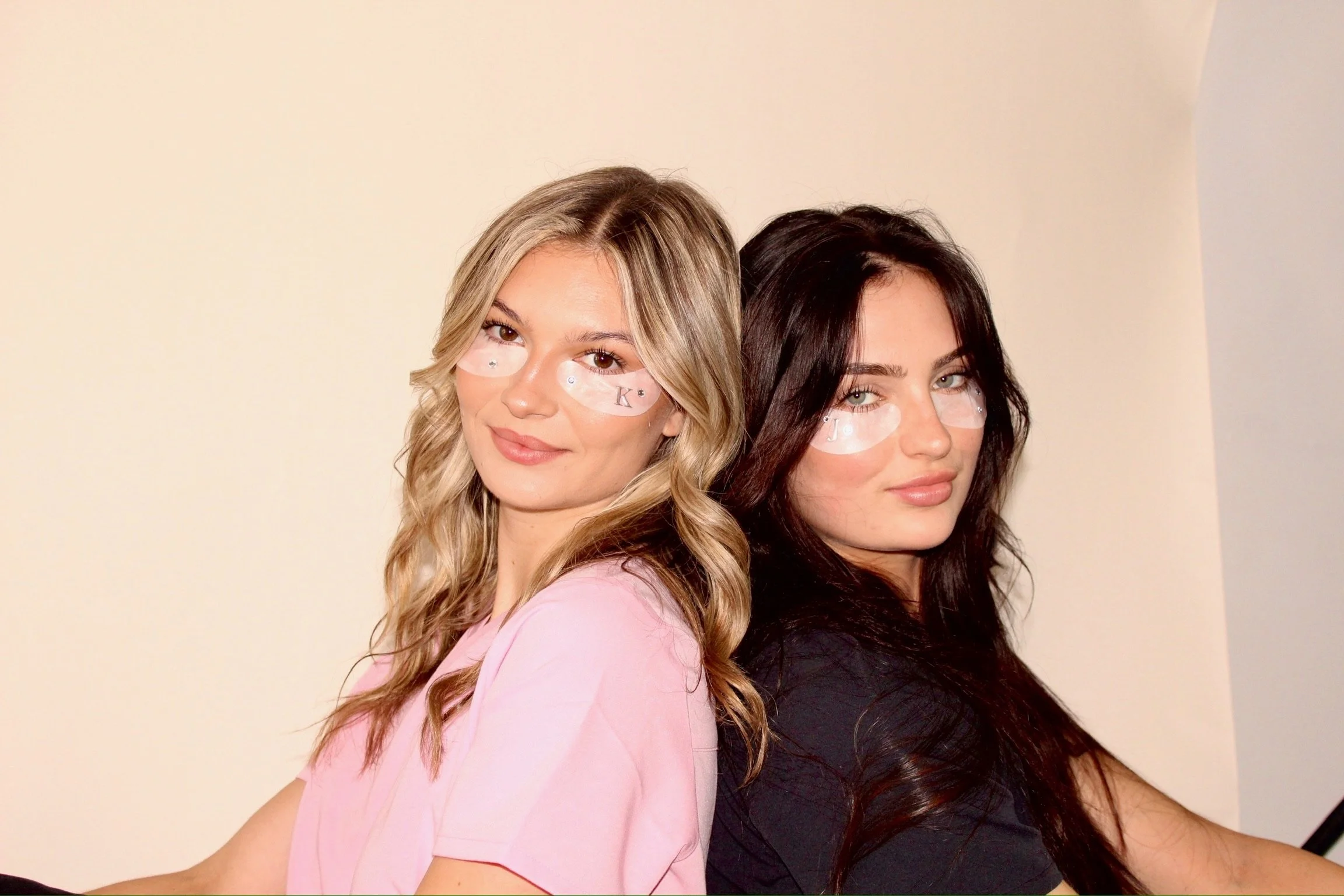 Two young women with long hair, one with blonde hair and the other with dark hair, standing back-to-back against a plain wall. They are wearing eye patches with small letters on them and appear to be posing for a photo.