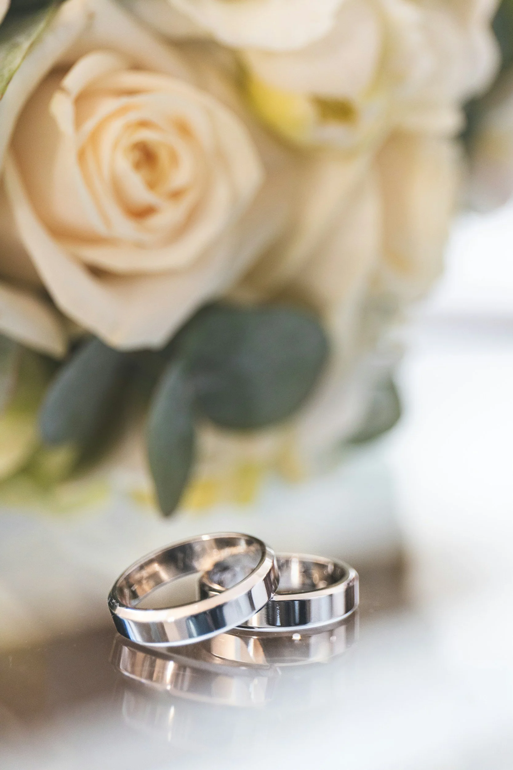 Close-up of two silver wedding bands on a reflective surface with blurred white roses and green leaves in the background.