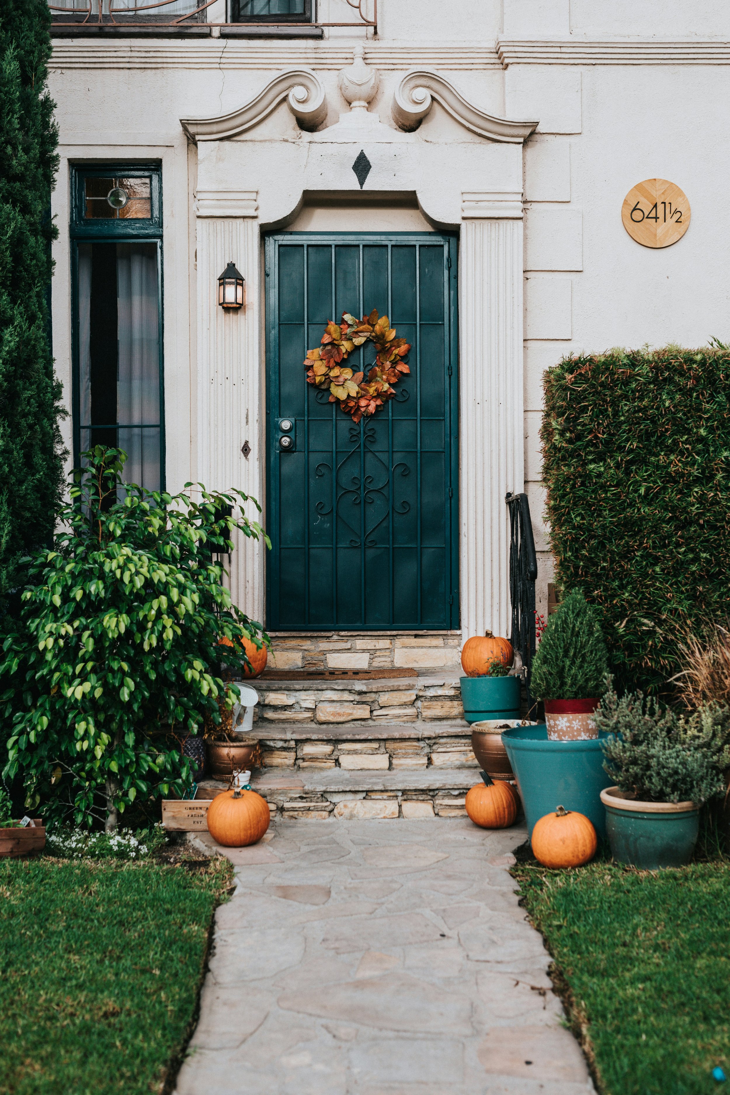 Home entrance with stone steps, decorated with pumpkins, potted plants, and a heart-shaped fall wreath on the door. The house has a white exterior, black iron gate, and a round house number plaque reading 641/2. The environment includes green bushes and a sidewalk.