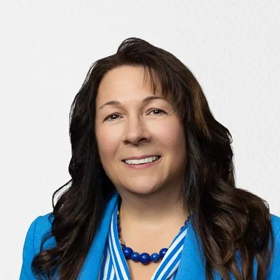 Portrait of a woman with long dark hair wearing a blue blazer and a blue beaded necklace, smiling against a white background.
