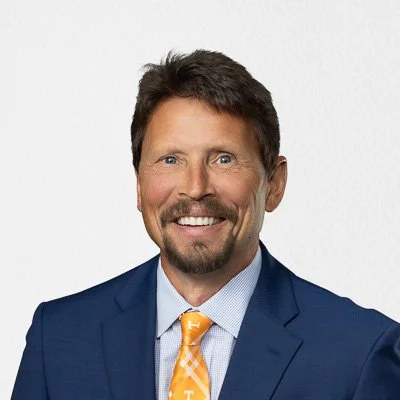 A man with dark hair, a beard, and a mustache wearing a blue suit, light blue shirt, and orange tie, smiling against a plain white background.