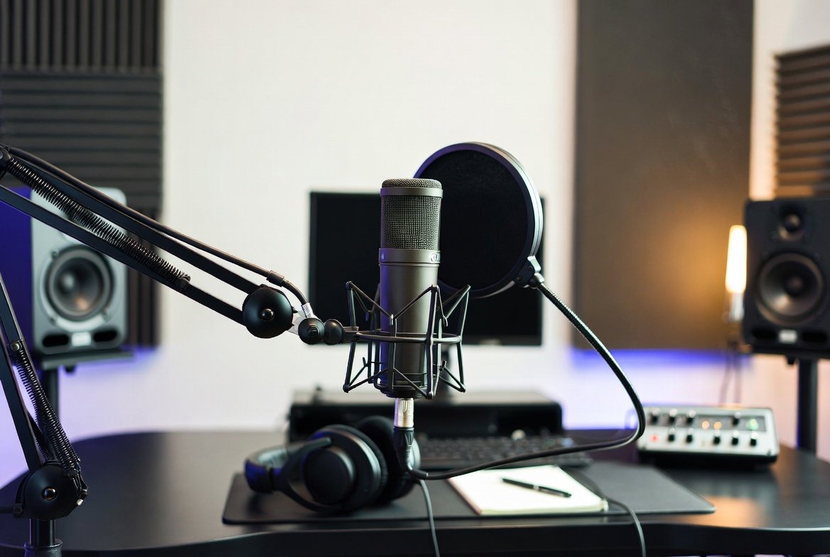 Recording studio setup with a microphone on a boom arm, headphones, studio speakers, a computer monitor, a control panel, and a notepad with a pen.