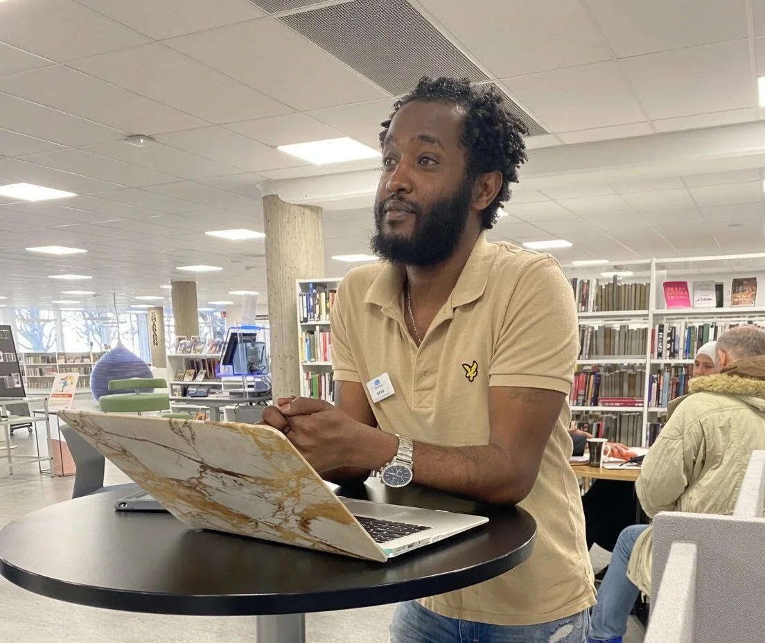 A man with a beard and curly hair sitting at a round table in a library, working on a laptop with a marble-patterned cover, wearing a beige polo shirt and a watch.