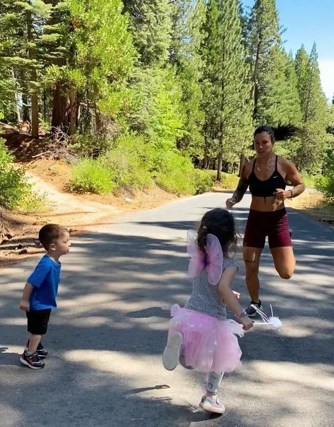 A woman is running on a forested road while two children, a boy and a girl dressed as a fairy, watch her.