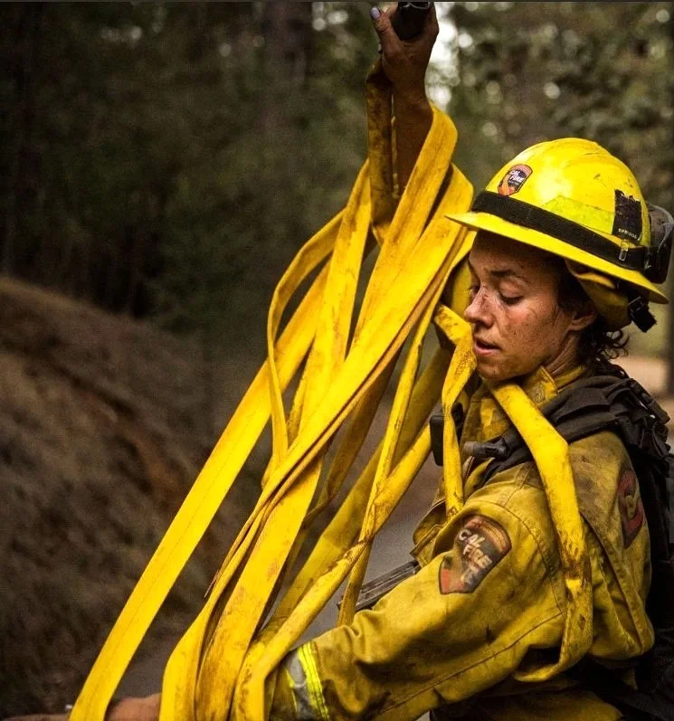 A firefighter in yellow gear and helmet, holding yellow fire hoses, standing in a forested area.