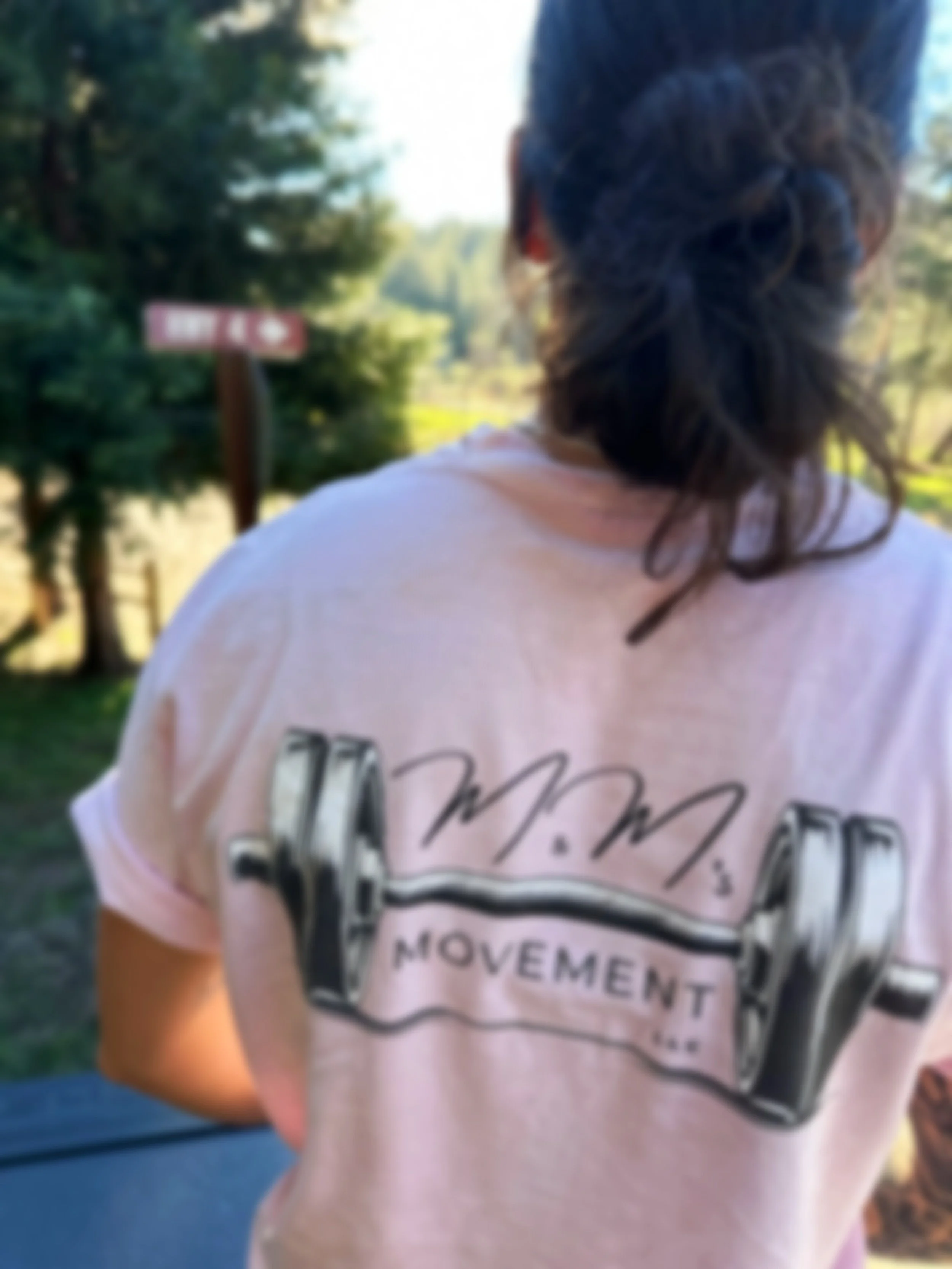 A person with braided hair wearing a pink T-shirt with a 'Me Movement' logo on the back, standing outdoors with trees and a sign in the background.