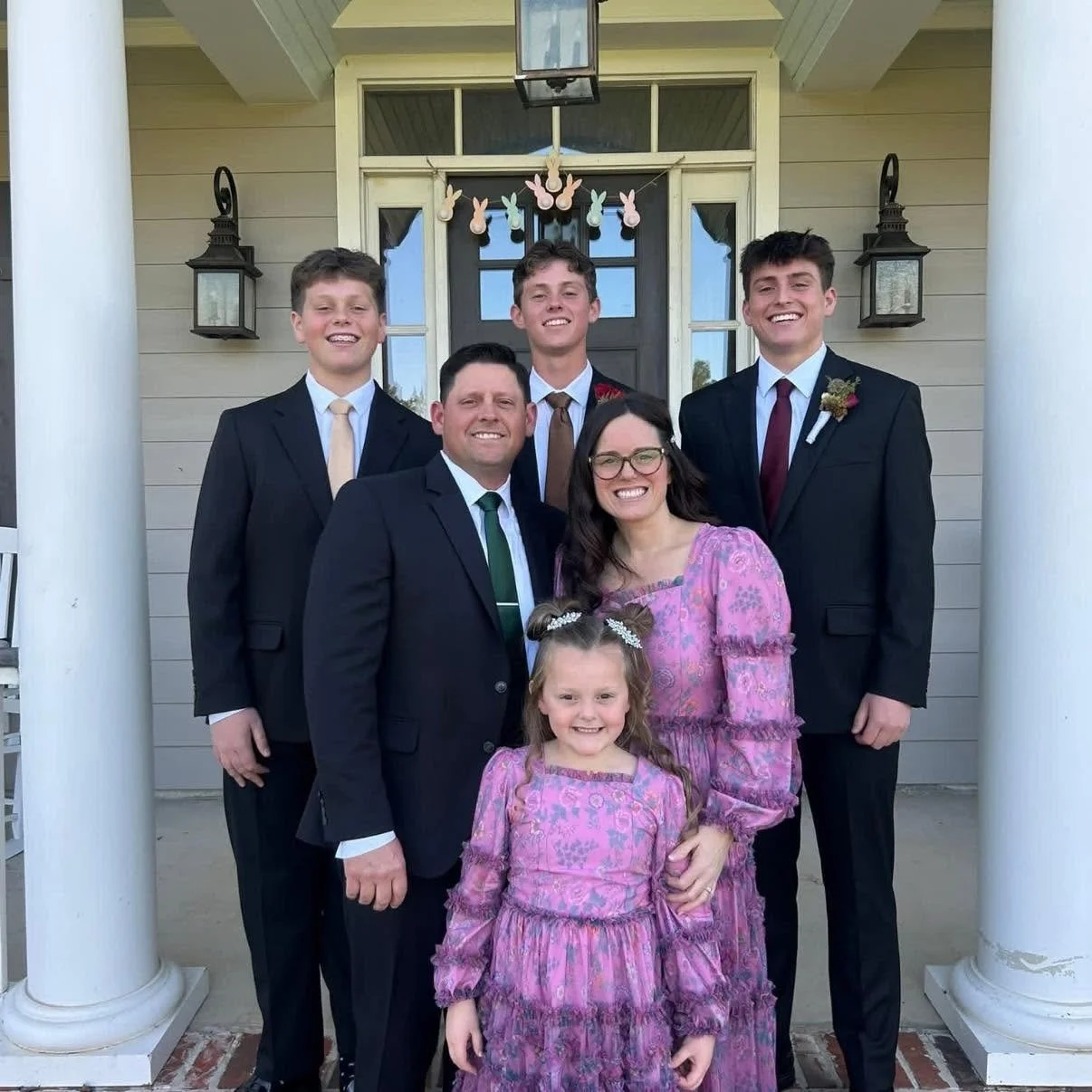 A group of six family members standing on the porch of a house, smiling for a photo. The group includes an adult man and woman, and four young men and a young girl, all dressed in formal attire. Decorated with pink and white Easter-themed bunny garland hanging above the door.