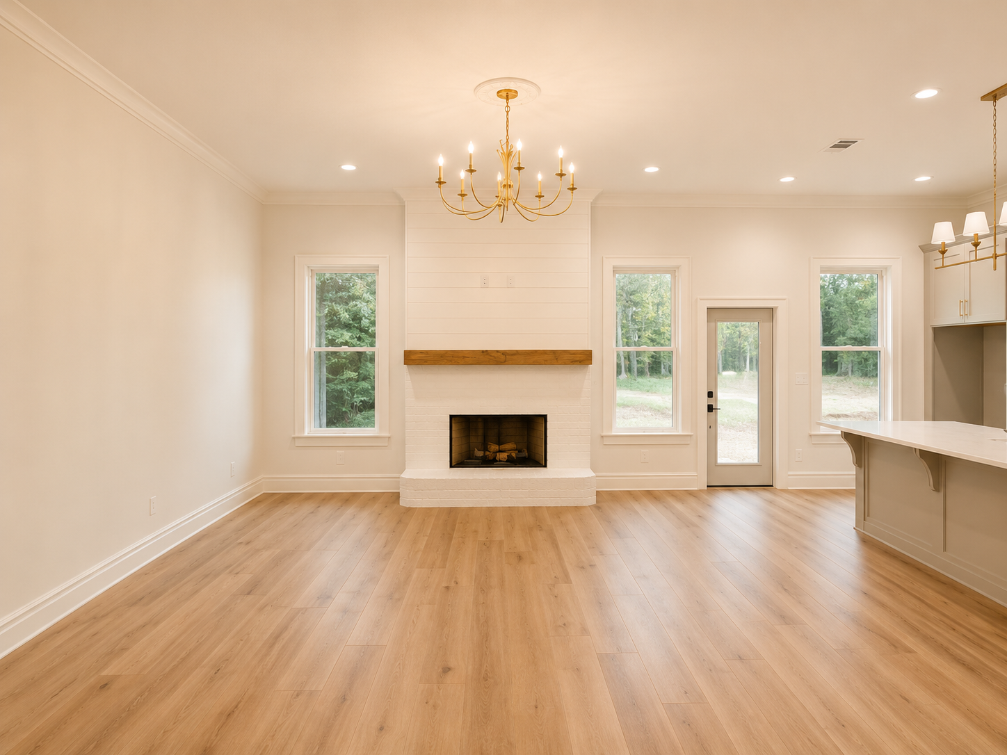 Living room with hardwood floors, white walls, fireplace with wooden mantel, two large windows, and a door leading outside. Ceiling lights and a chandelier hang from the ceiling.