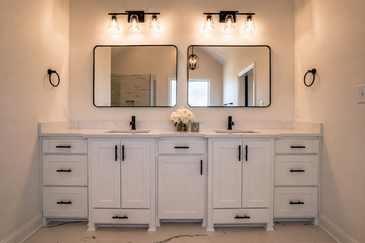 White bathroom vanity with two sinks, black fixtures, and two large mirrors, decorated with a vase of white flowers and framed art, lit by modern black light fixtures and wall-mounted towel rings.