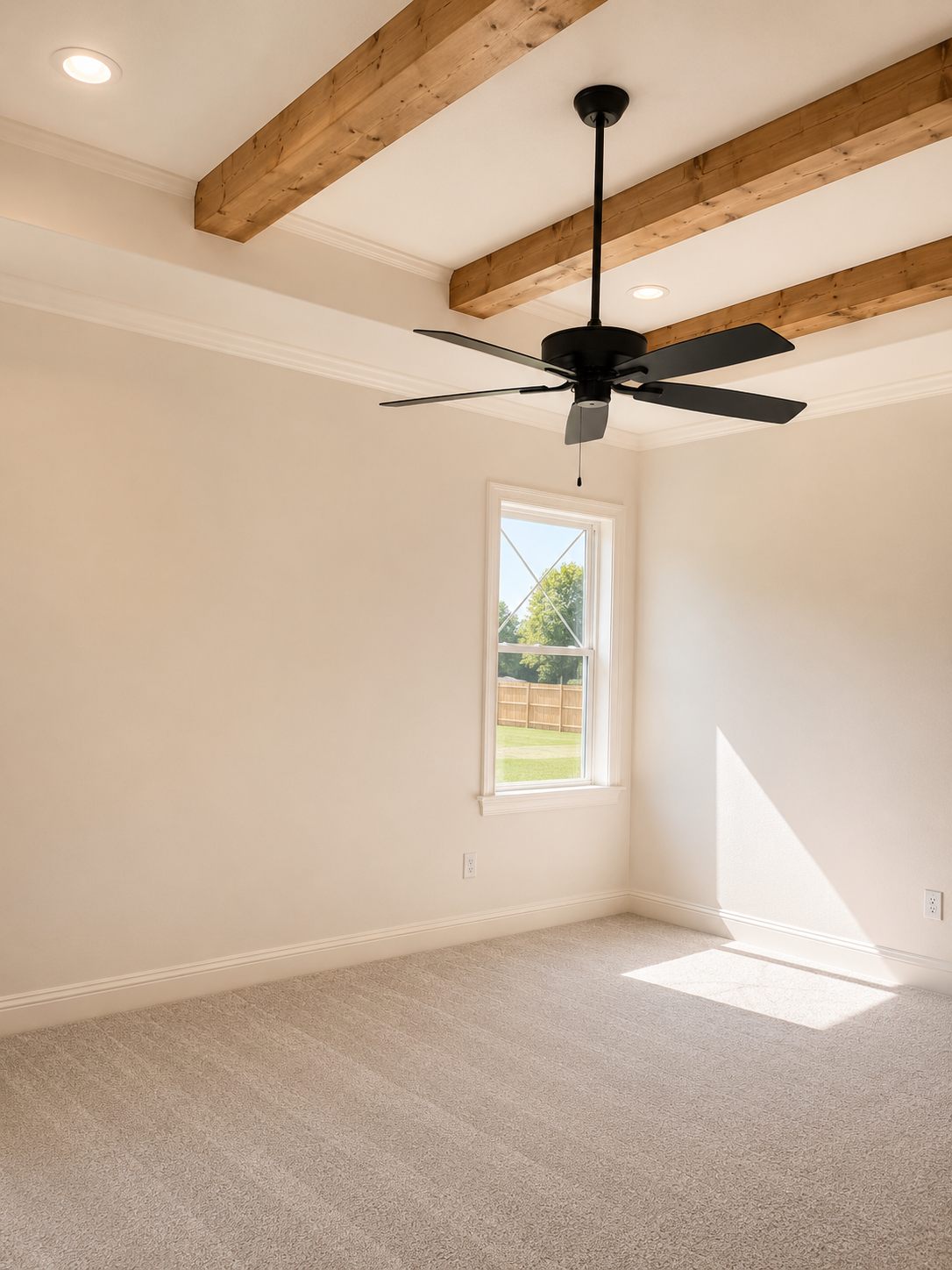 Empty room with beige carpet, white walls, a window showing a fenced backyard, a ceiling fan, recessed lighting, and wood ceiling beams.