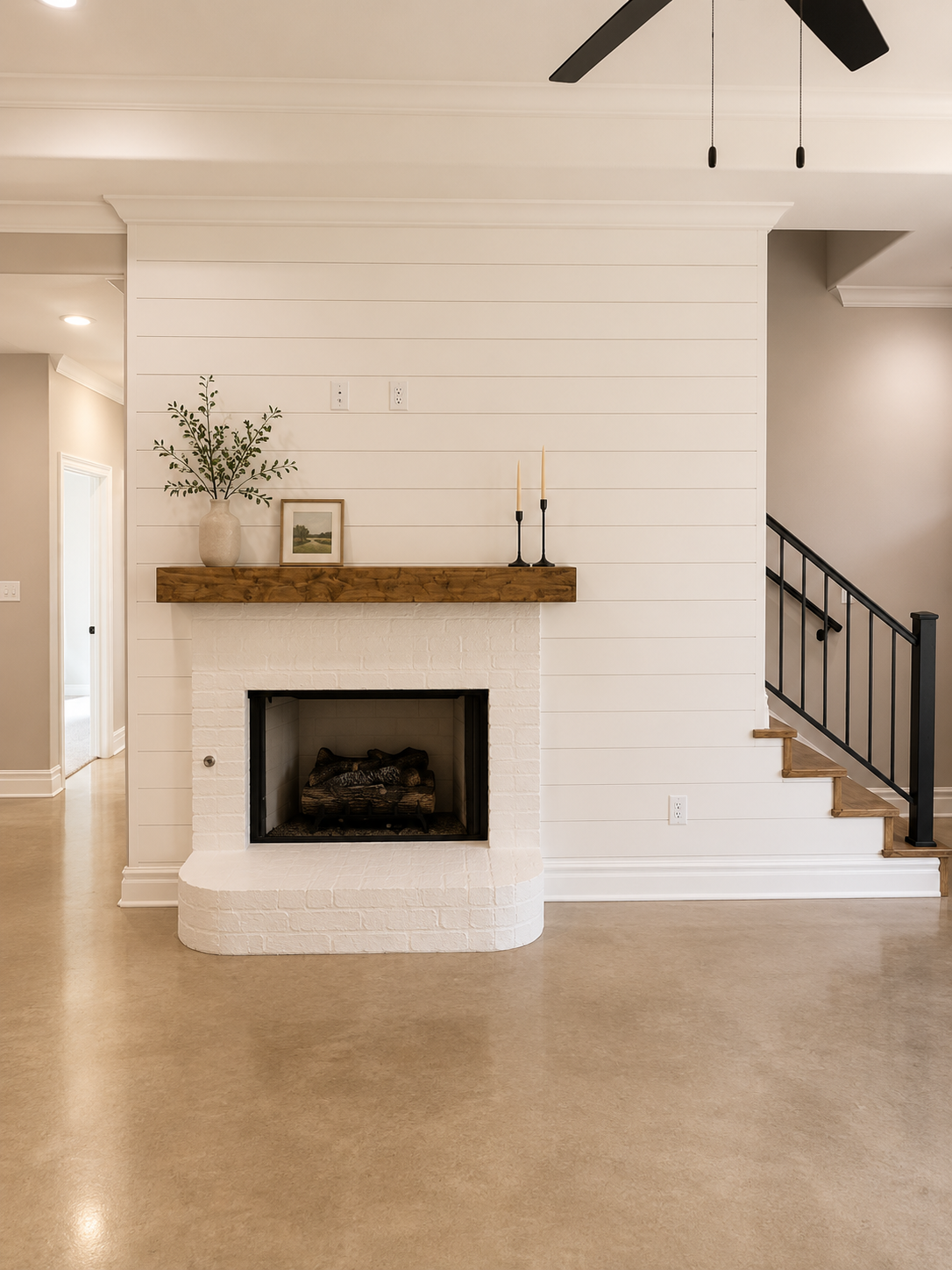 Living room with a white brick fireplace, wooden mantel, a decorative vase with greenery, framed artwork, and two candlesticks, with stairs on the right and a ceiling fan above.