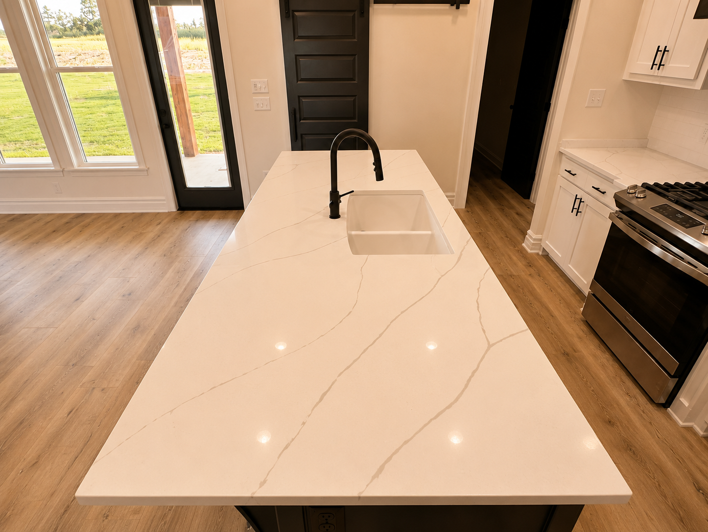 Kitchen island with a white marble countertop and black faucet, with a view of a patio door overlooking a green yard, white cabinets, and stainless steel oven.