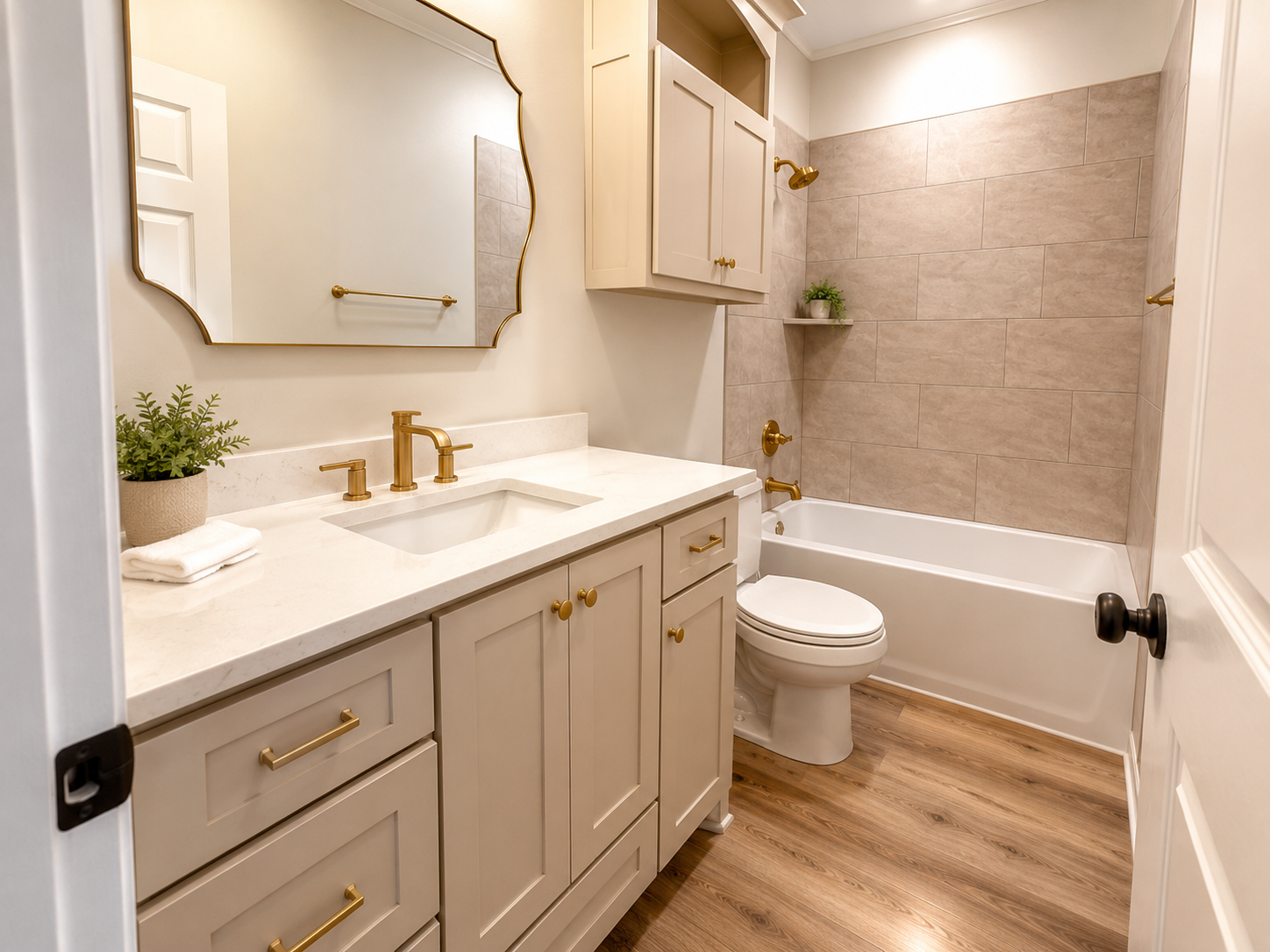 A clean, modern bathroom with cream-colored cabinets, a white countertop, a brass faucet, large wall mirror, a toilet, a bathtub with a beige tiled wall, a small shelf with a green plant, and hardwood flooring.