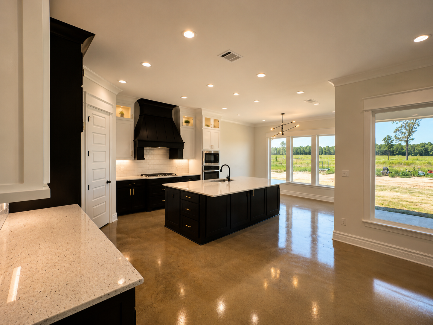 Modern kitchen with black cabinets and island, white countertops, large windows overlooking a field, and recessed lighting.