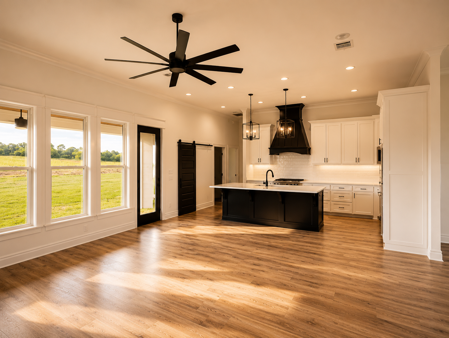 Empty modern kitchen with white cabinets, black range hood, black island, large windows overlooking a grassy field, wood flooring, and ceiling fan.