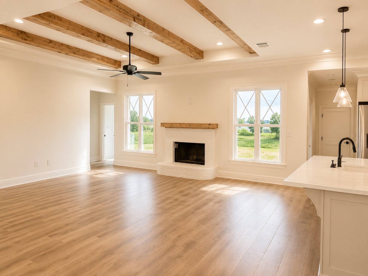 Empty living room with wood floors, white walls, a white brick fireplace with a wooden mantel, large double-pane windows showing a green outdoor landscape, a ceiling fan with black blades, and wood ceiling beams.