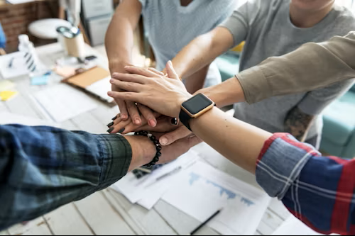 Group of people placing their hands together in a teamwork gesture in an office setting.