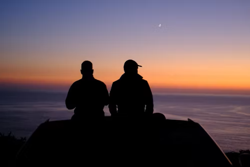 Two people sitting on a hilltop during sunset, overlooking the ocean with a crescent moon in the sky.