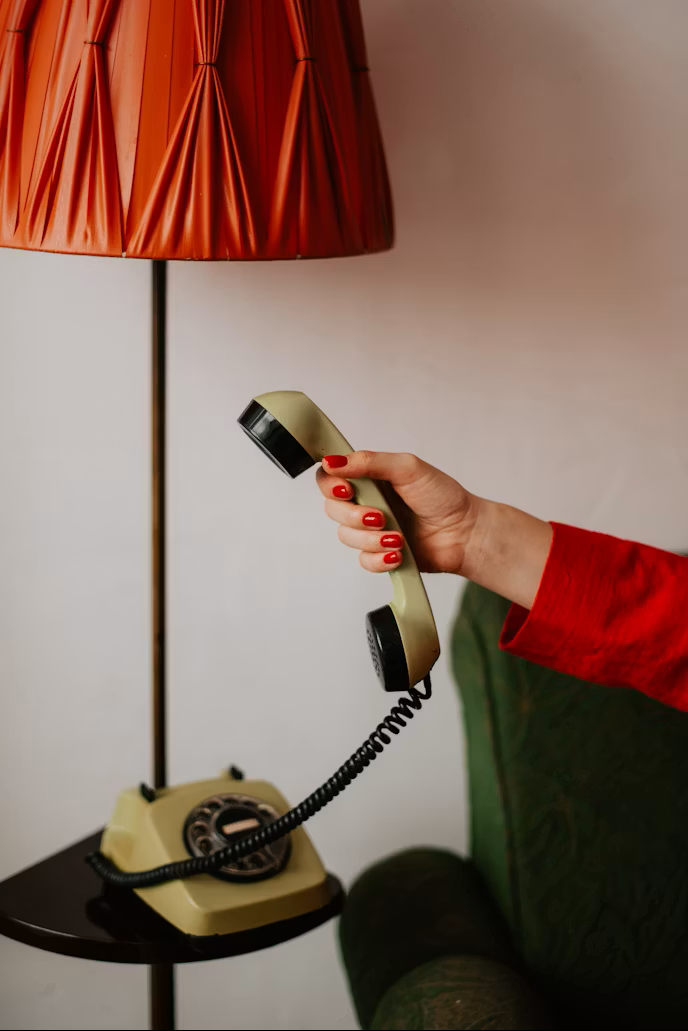 Person holding a vintage beige rotary phone corded telephone in front of a red lampshade and a white wall.