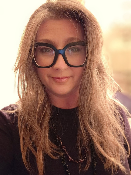Woman with shoulder-length light brown hair wearing large black glasses and a black top, smiling softly, outdoor background with warm lighting.