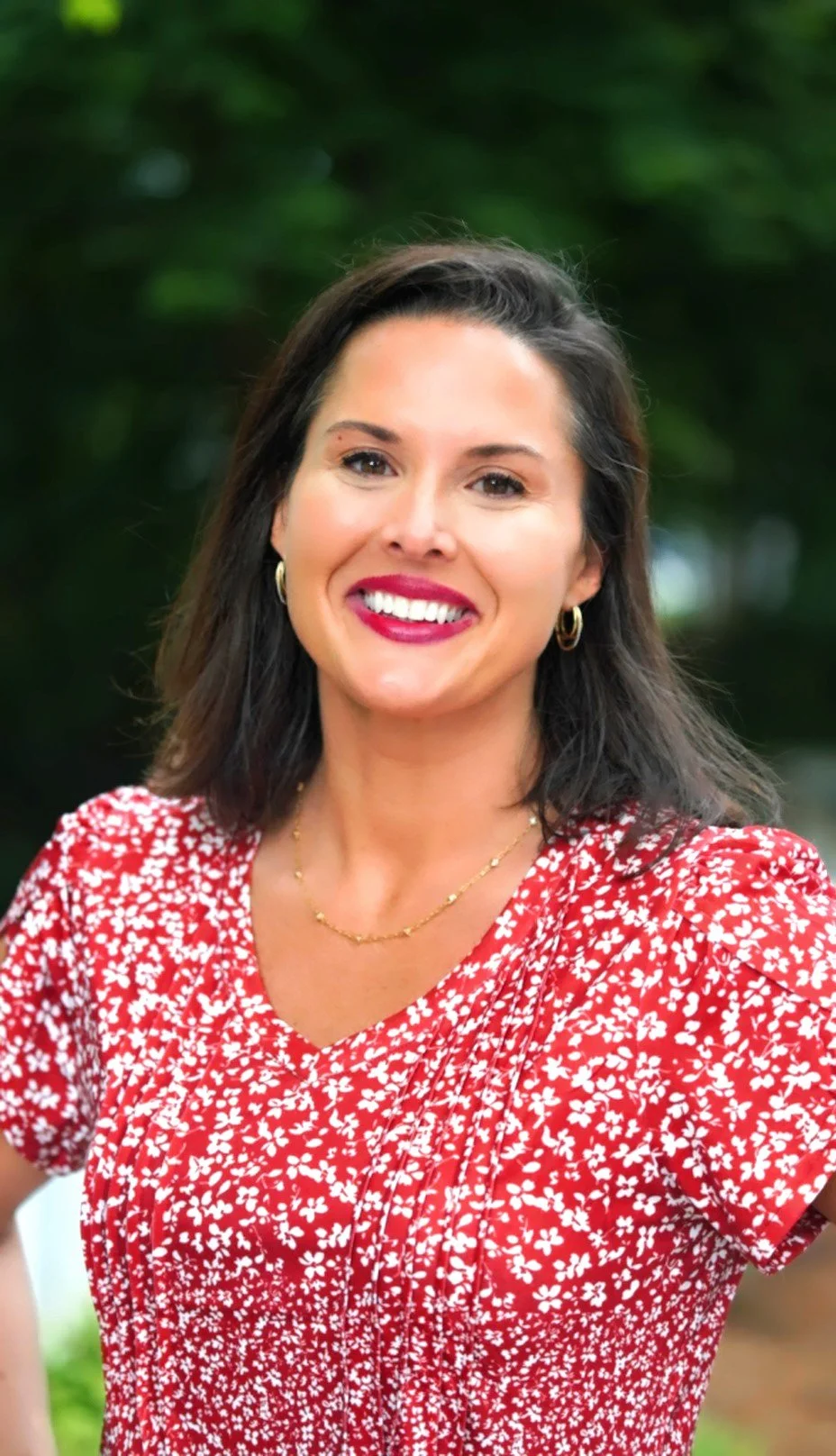 A woman smiling with dark shoulder-length hair, wearing a red and white floral dress, gold hoop earrings, and a gold necklace, standing outdoors with blurred green foliage in the background.