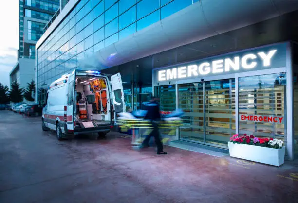 An ambulance parked outside an emergency entrance with a nurse or paramedic pushing a stretcher into the hospital.