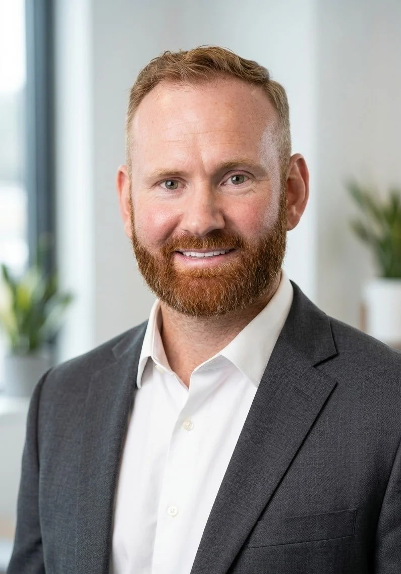 Headshot of a smiling man with red hair and beard wearing a dark suit and white shirt, standing in an office with plants in the background.