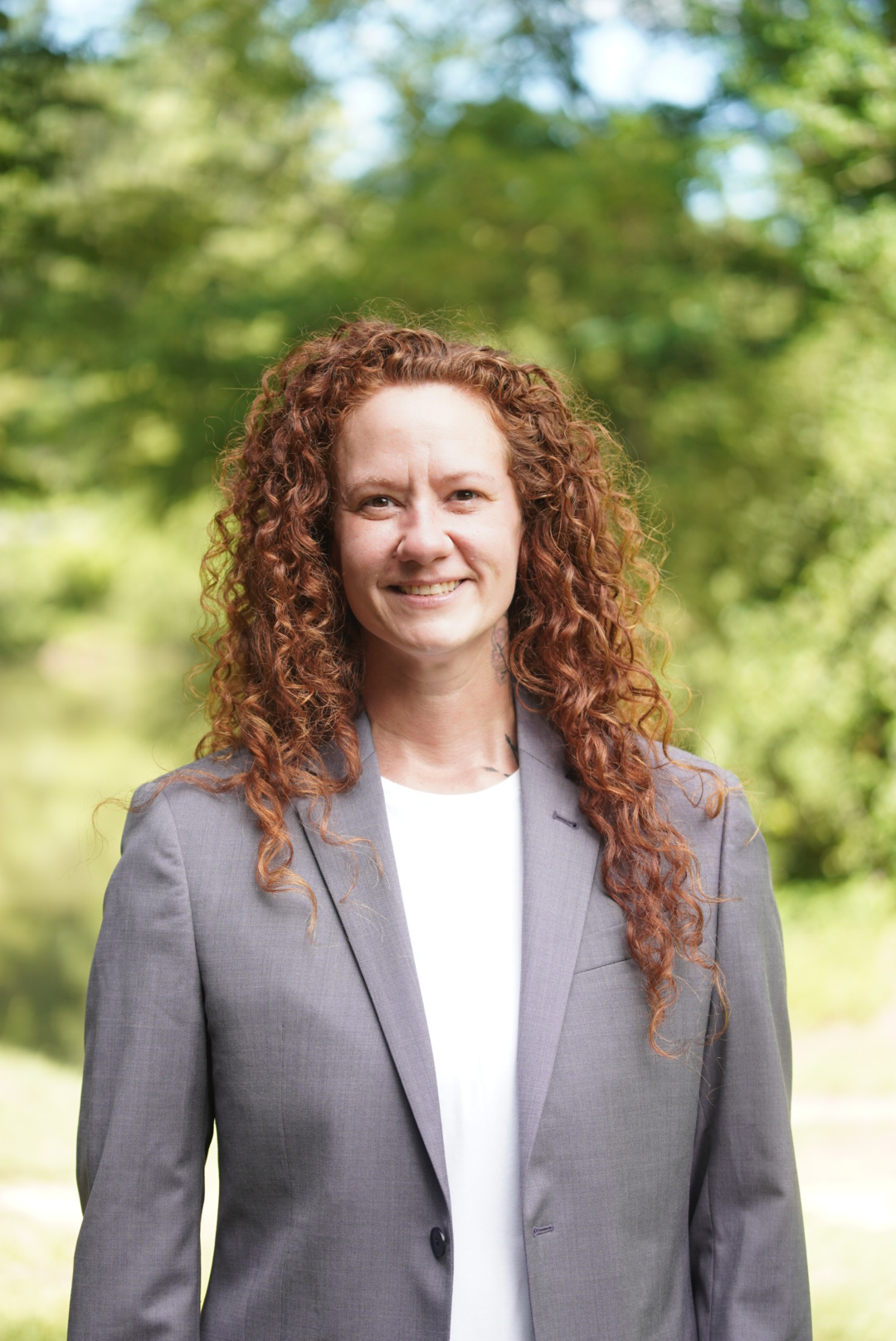 A woman with curly red hair standing outdoors in front of green trees, wearing a gray blazer over a white top, smiling at the camera.