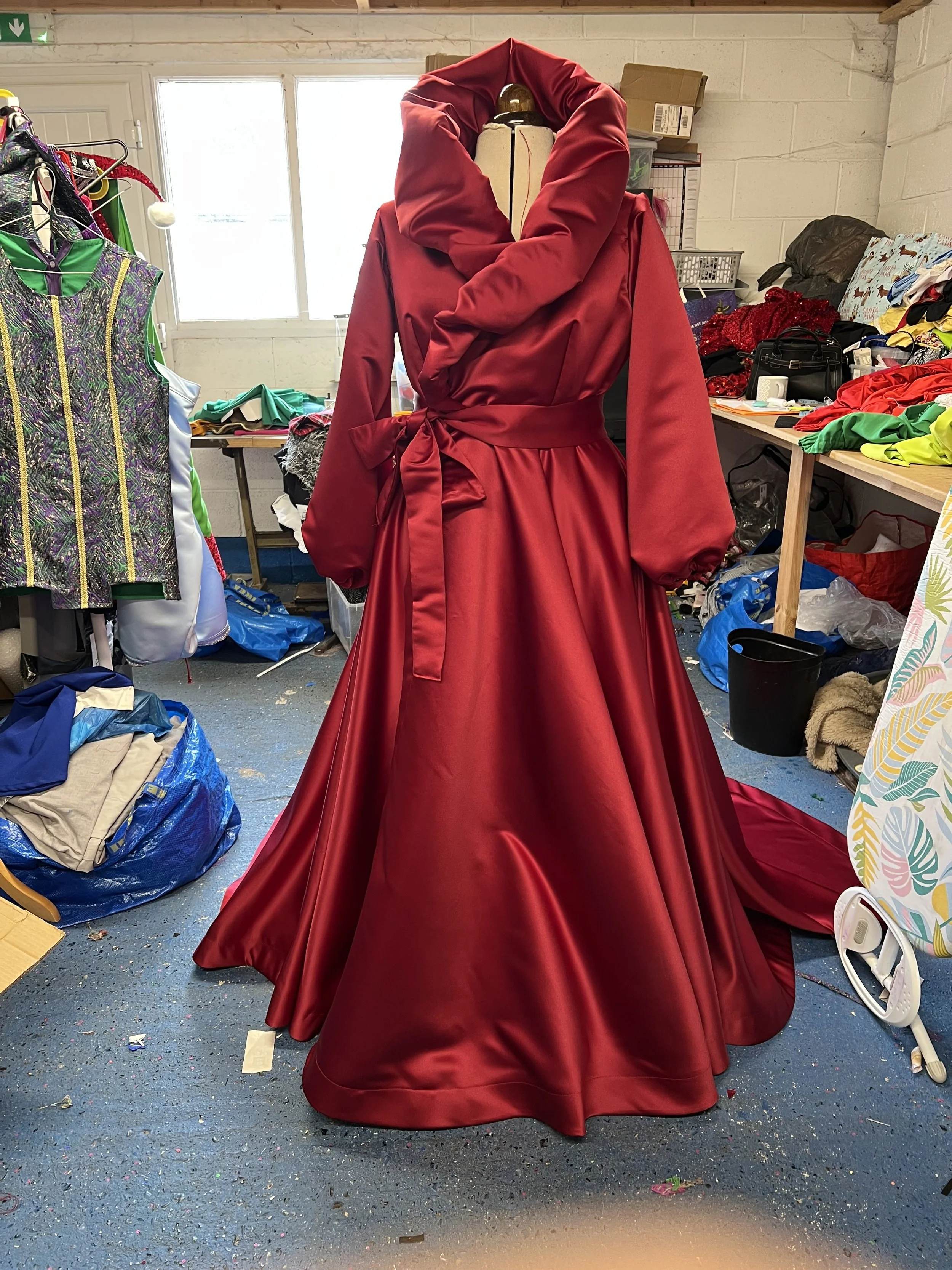 A red, voluminous dress on a mannequin in a cluttered room, with fabric, clothing, and miscellaneous items scattered around.