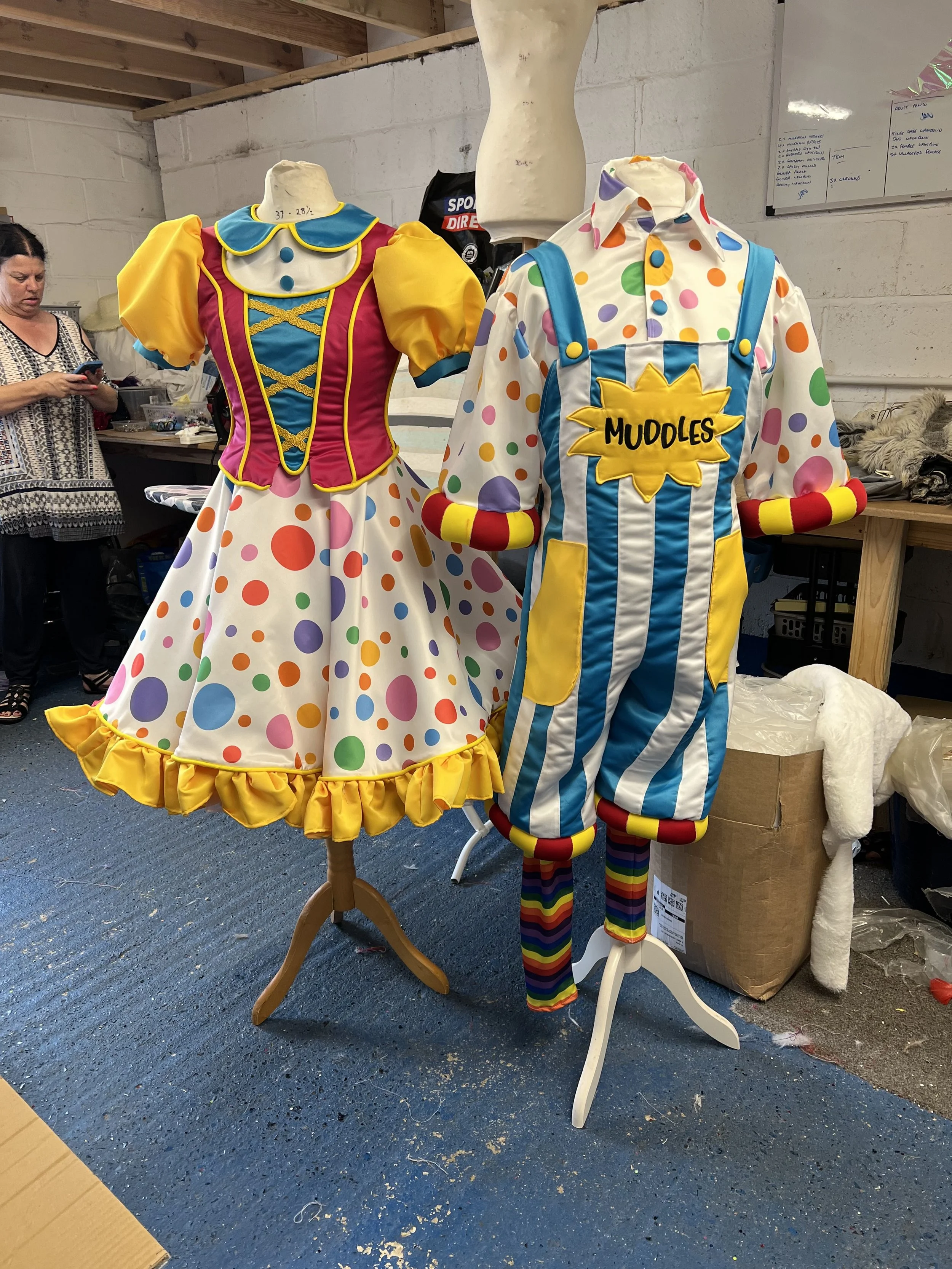Colorful clown costumes on mannequins in a storage or workshop space. One costume has a multicolored polka-dot pattern, and the other has a striped pattern with a large yellow patch labeled 'MUDDLES.' A woman is visible in the background looking at h