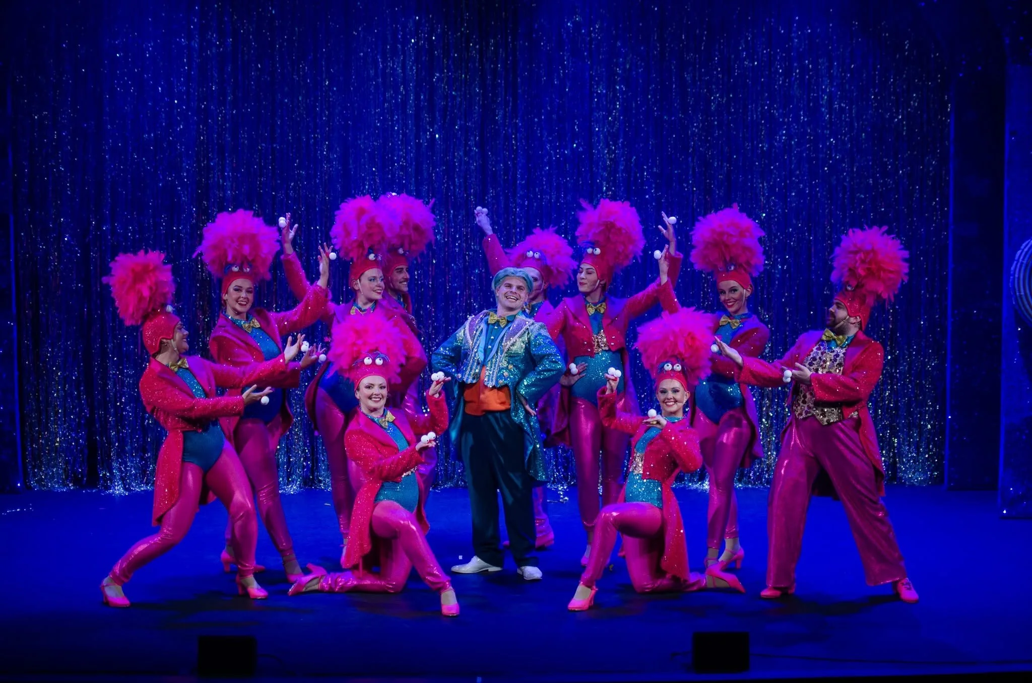 A group of performers dressed in colorful costumes with pink, fluffy headdresses, and blue and pink outfits, dancing on stage with a glittering blue curtain backdrop.