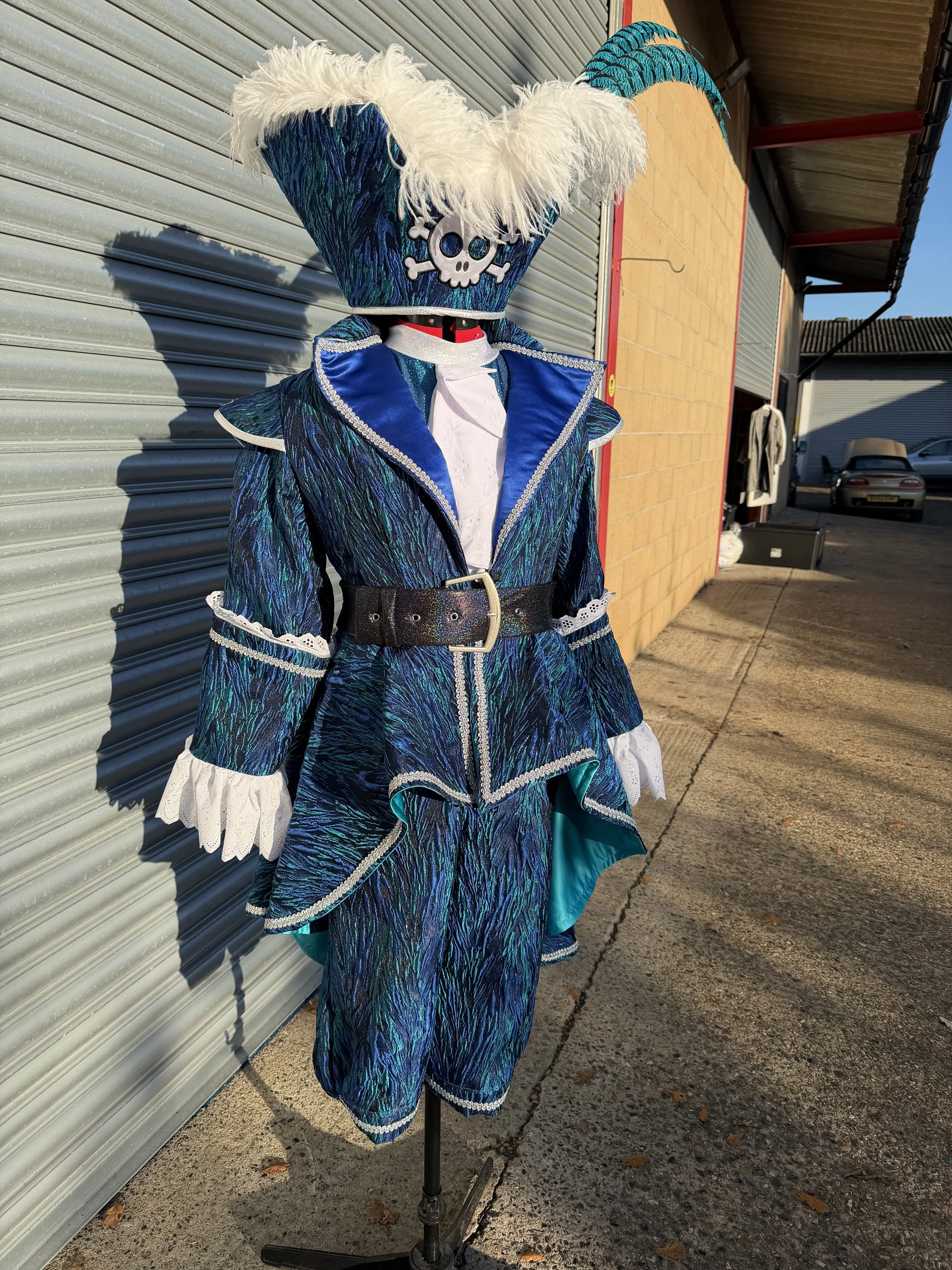 A costume resembling a character from a pirate or fantasy theme, with a large headpiece featuring a skull and crossbones, decorated with white feathers, shiny blue and teal fabric, and lace details. The costume is displayed on a mannequin outside nea