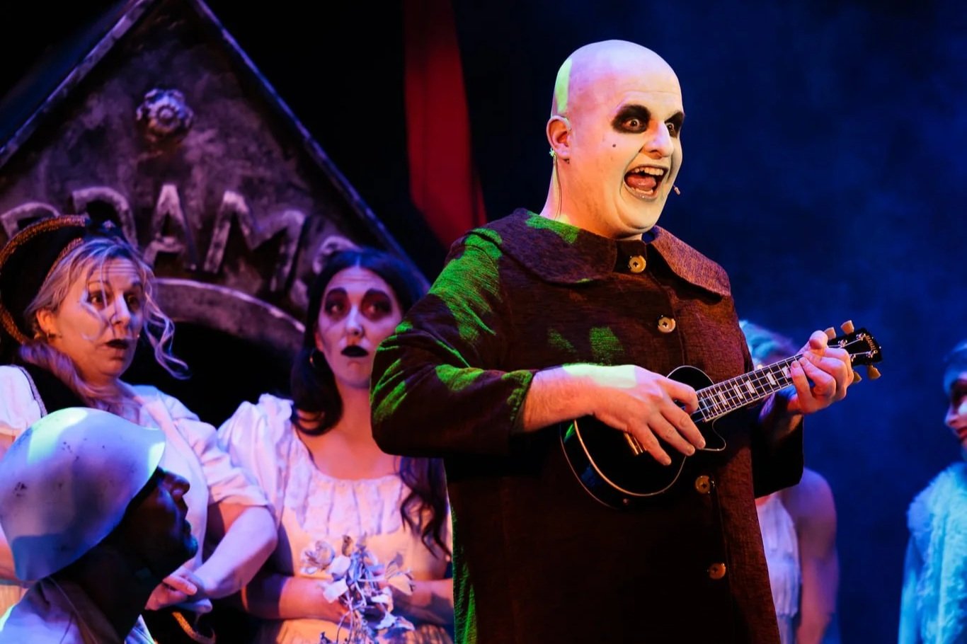 Actors in a theatrical performance, with one dressed as a spooky clown with a bald head and dark makeup, playing a small guitar, while others in costume look surprised or frightened, set against a dark stage background.