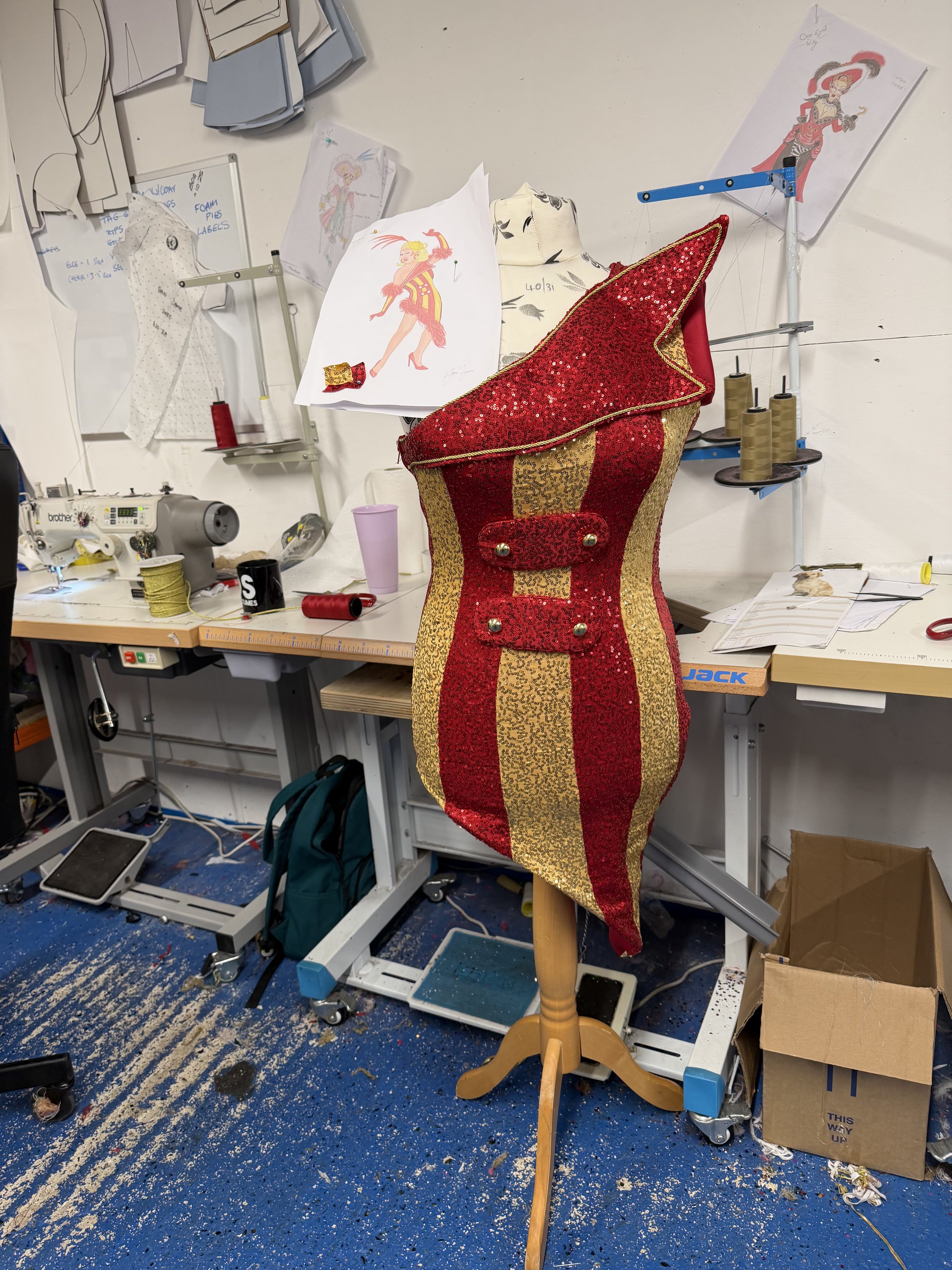 A workspace with a mannequin draped in a gold and red sequined costume, surrounded by sewing supplies, sketches, and fabric on a cluttered table and wall.