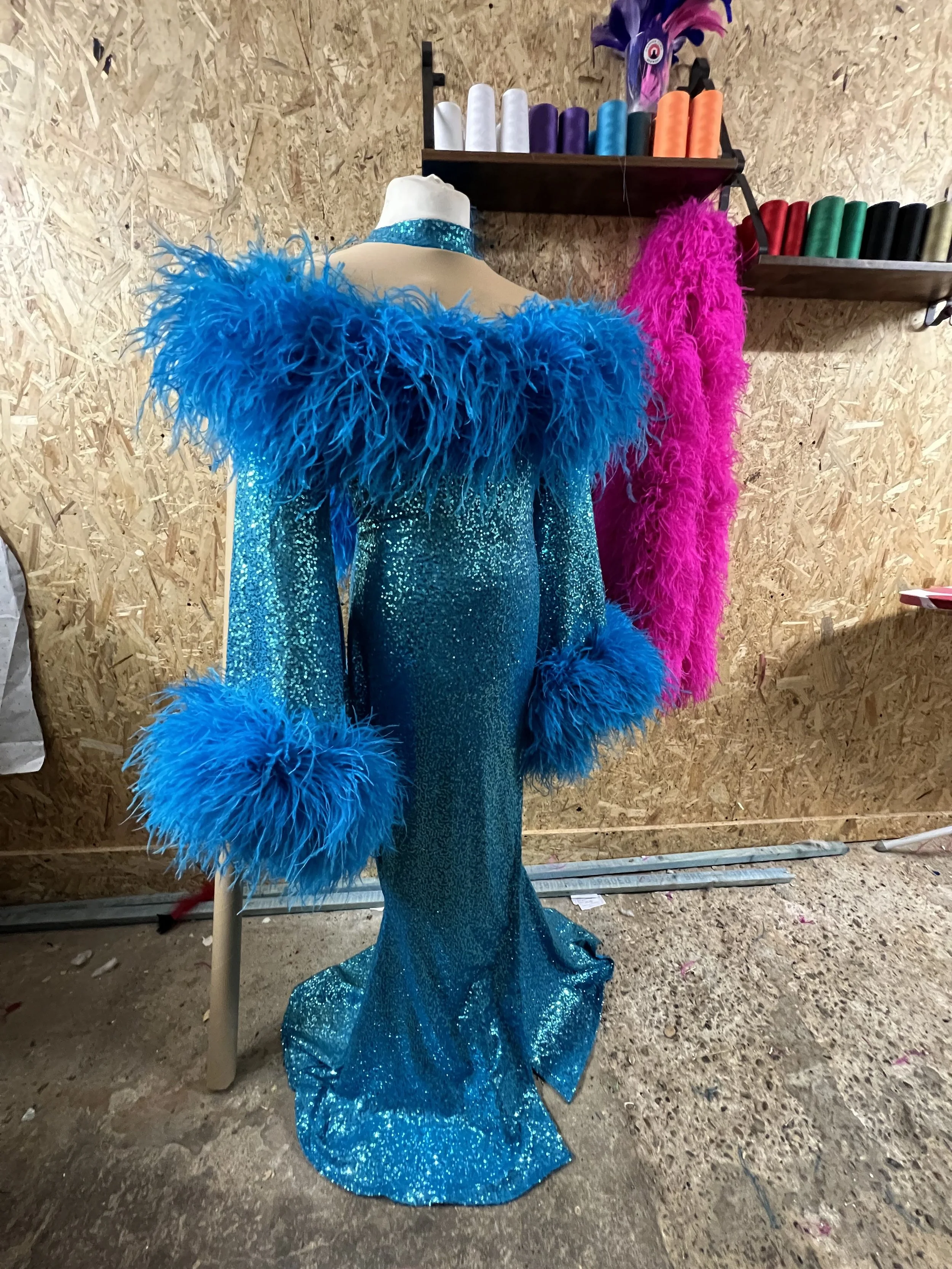 Blue and pink feathered costume with sequined fabric on a mannequin in a workshop with shelves of colorful spools of thread.