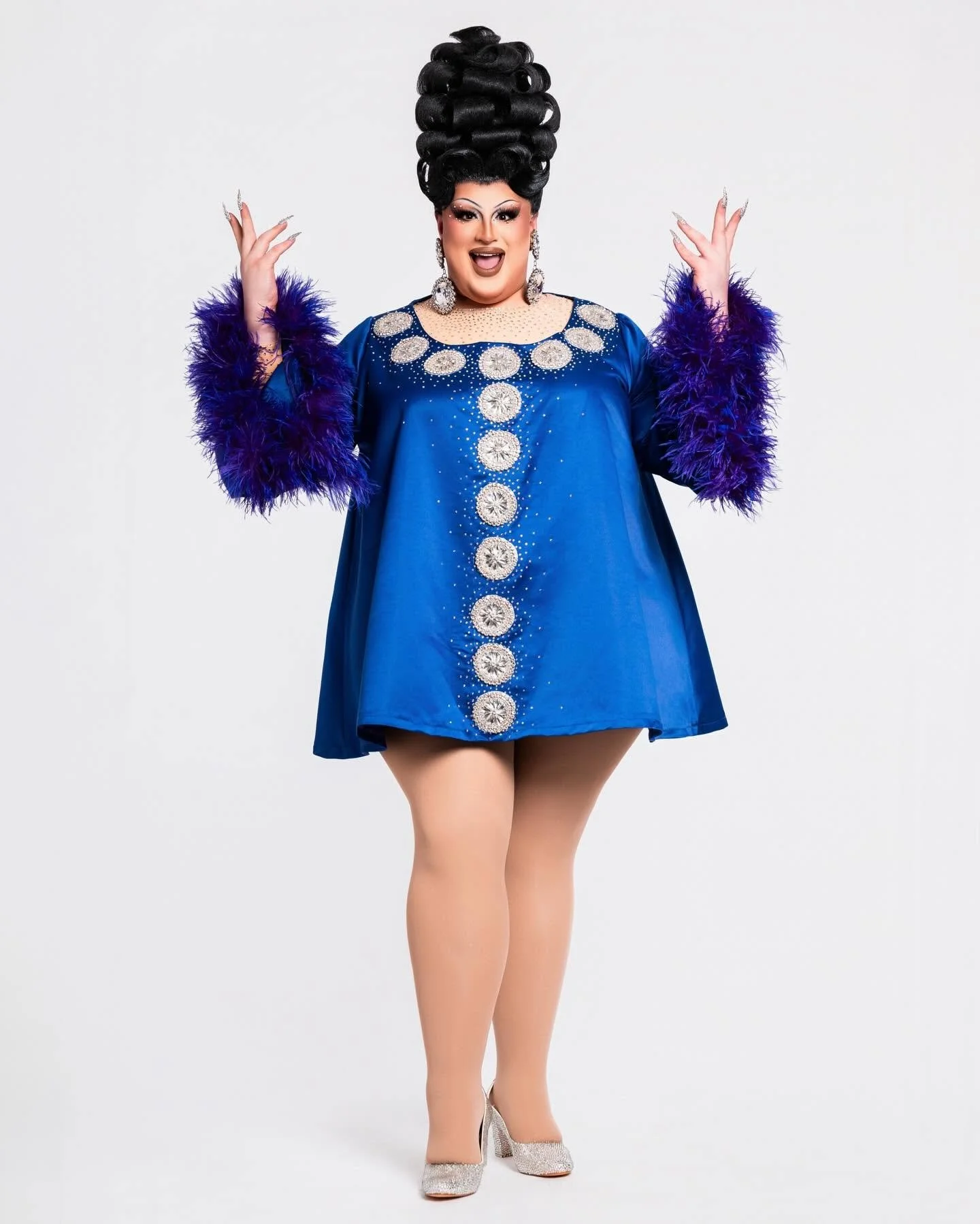 A person dressed in a blue dress with silver embellishments and purple feathered sleeves, platinum high heels, and large earrings, with styled black hair in an elaborate updo, smiling with arms raised against a plain white background.