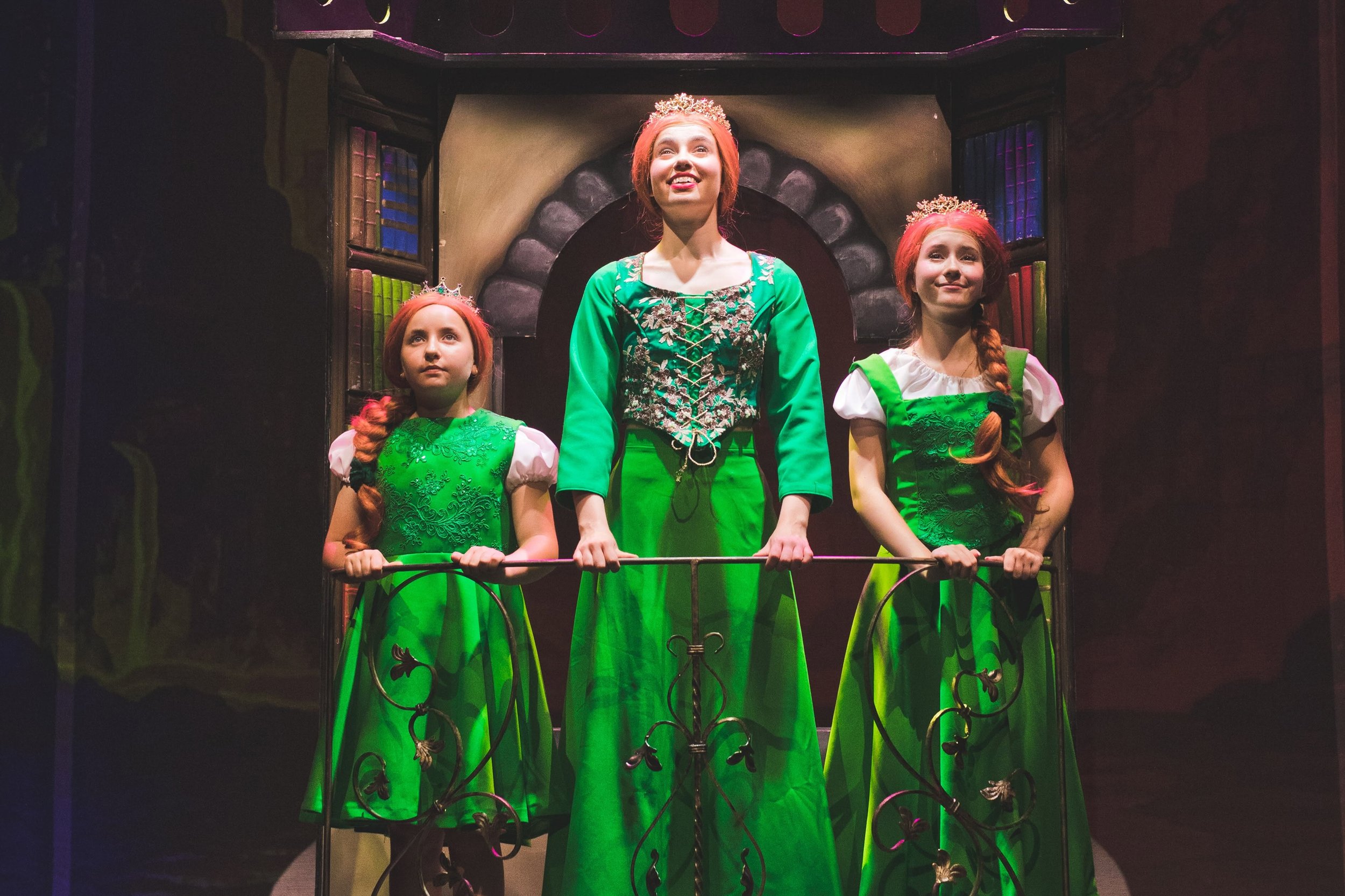 Three women dressed as princesses standing on a stage, with ornate green dresses, tiaras, and long braided hair, in front of a dark, castle-like backdrop.
