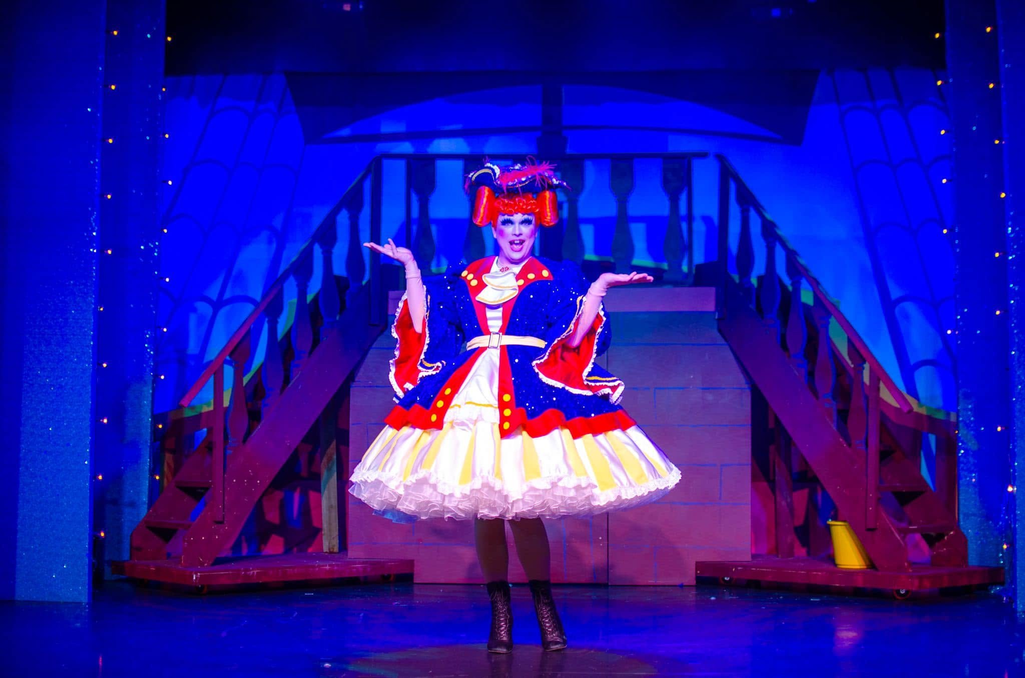 Performer dressed as a clown in colorful costume on stage, with a blue background and a staircase in the back.
