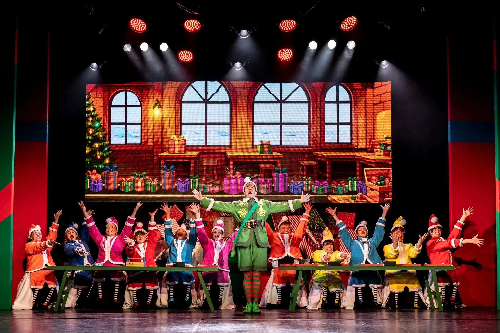 Group of performers wearing colorful Christmas costumes on stage during a festive show, with a large digital backdrop depicting a holiday scene and decorated Christmas tree.